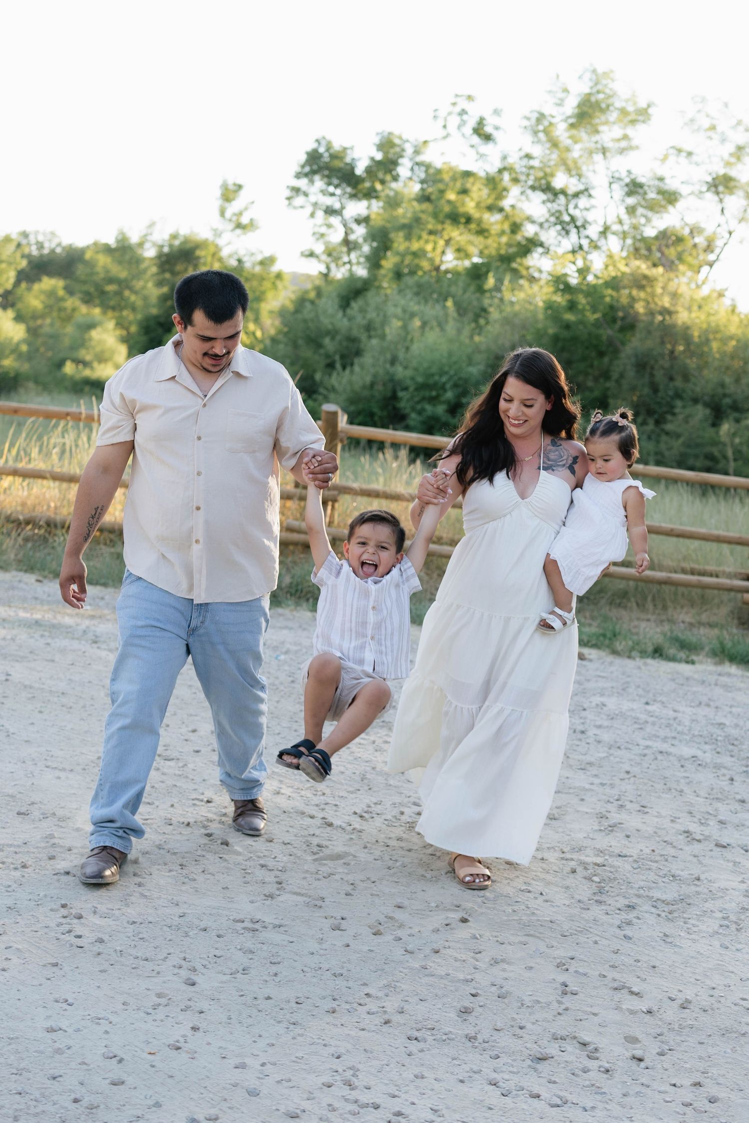 Happy family of four walking on a sandy path, parents swinging young boy while mother holds baby girl outdoors.