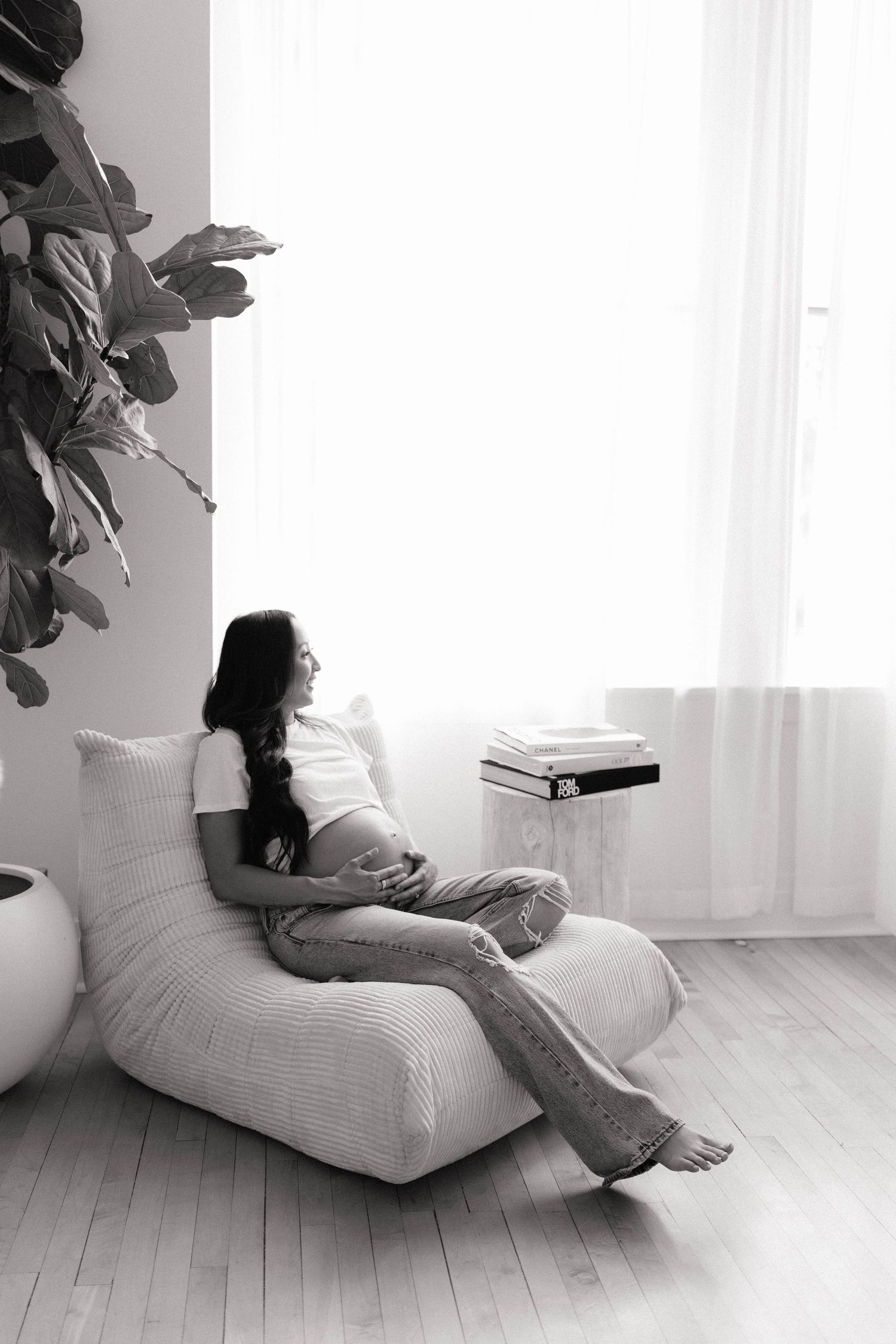 Black and white photo of a pregnant woman relaxing in a cozy chair by a bright window with a plant nearby.
