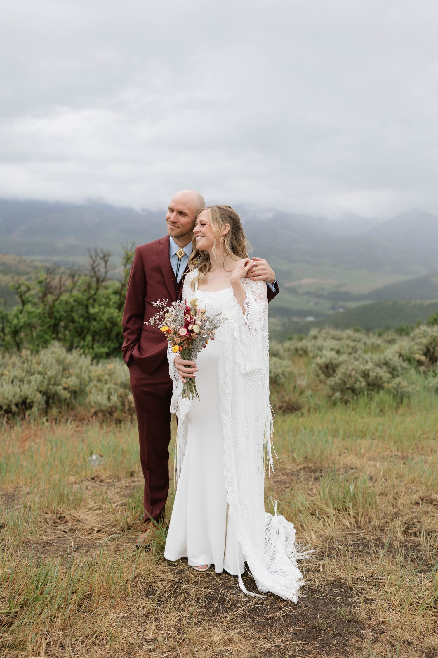 A bride in a white boho gown holds a wildflower bouquet beside a groom in a burgundy suit with mountain views.