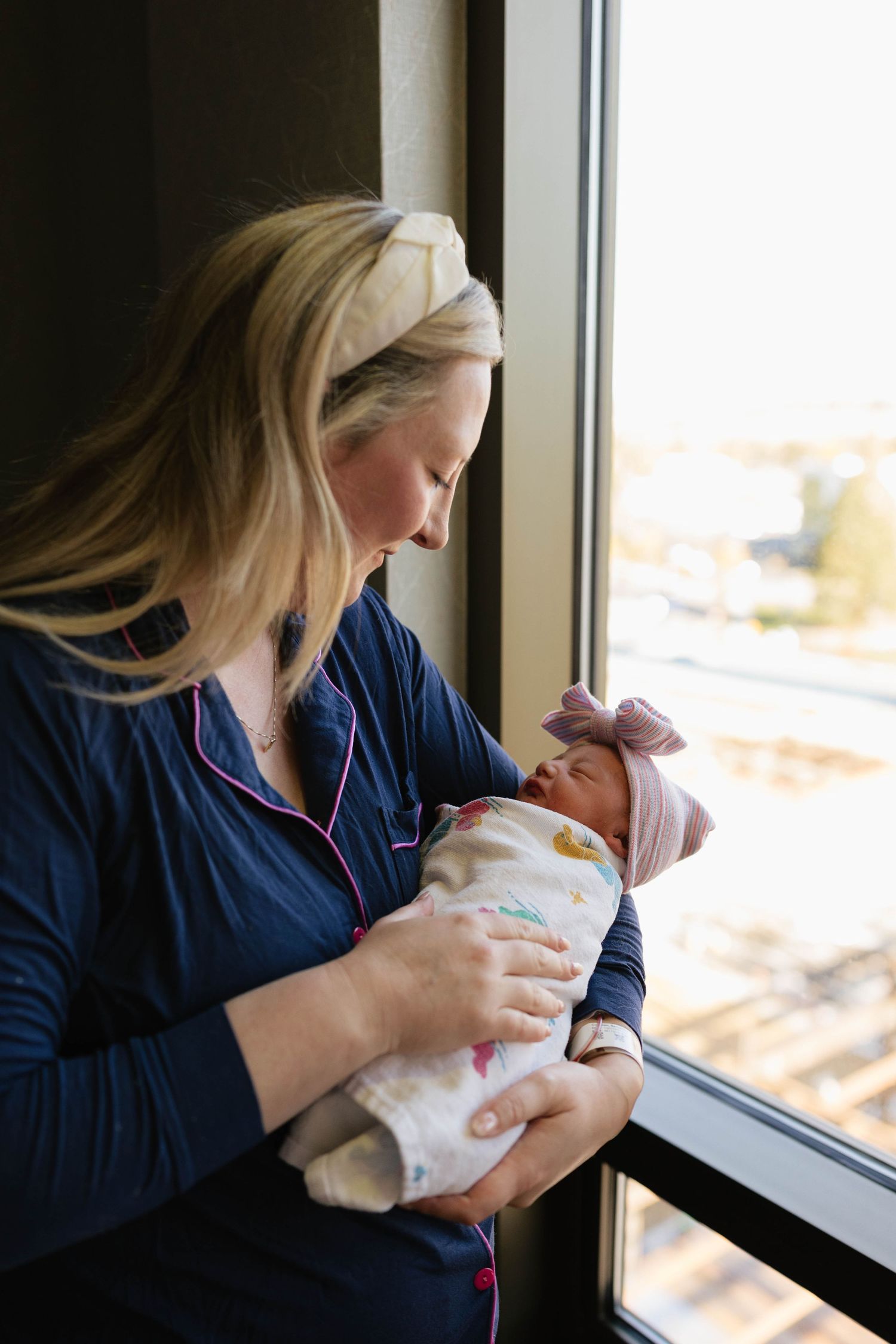 Woman in navy robe holding newborn by window in hospital room.