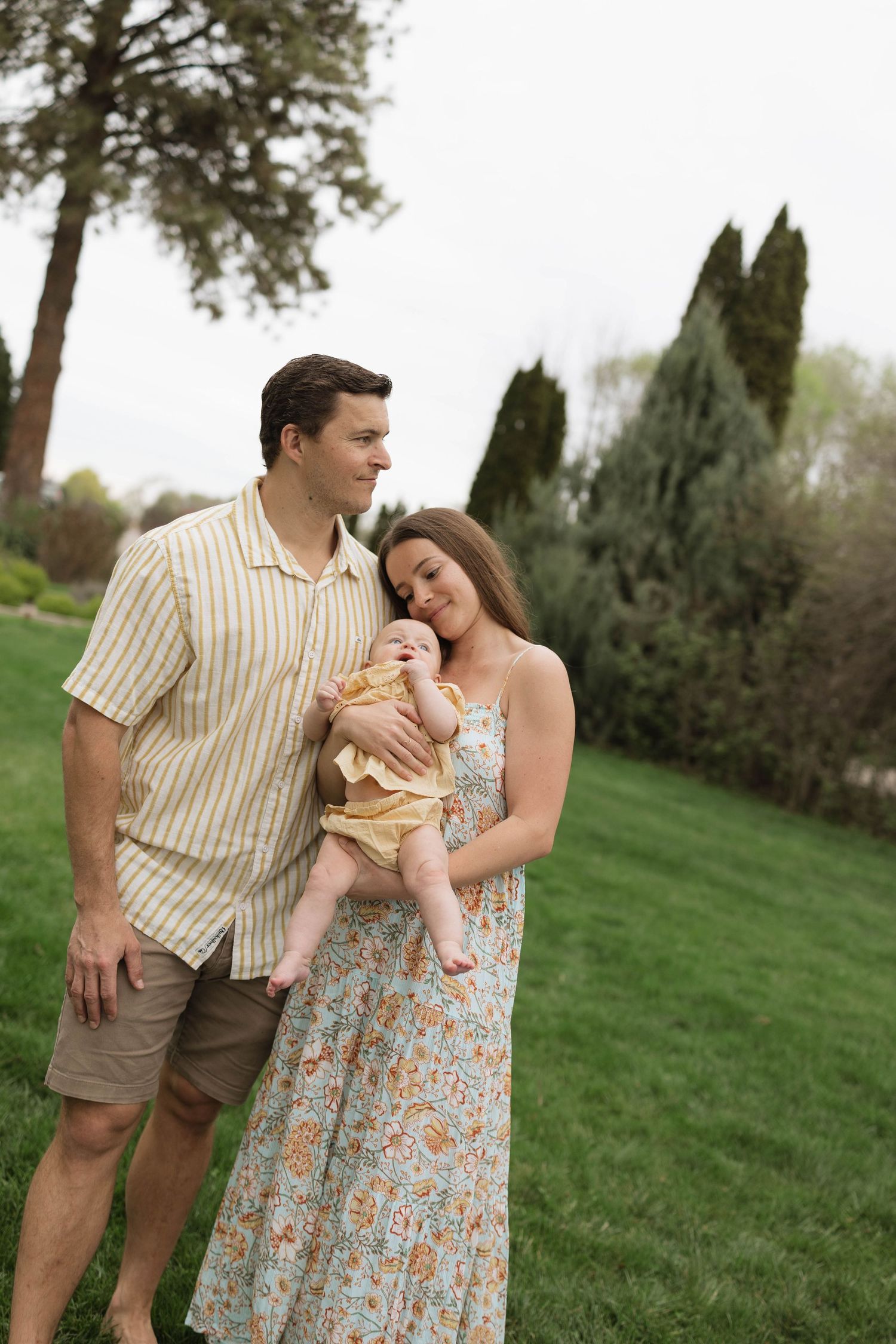 A couple stands together on a grassy lawn with their baby, wearing casual spring attire against a backdrop of evergreen trees.