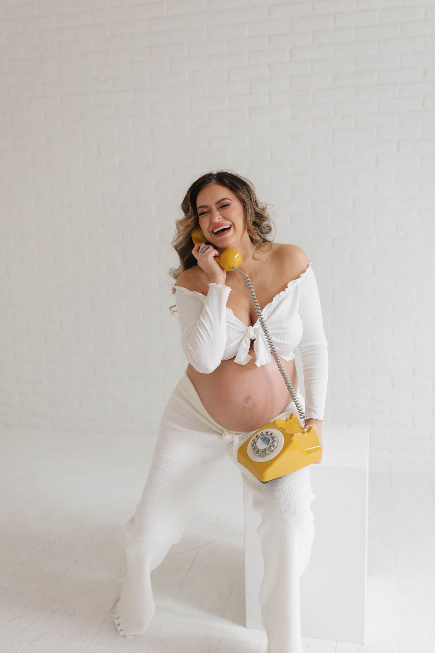 A pregnant woman in white attire laughs while holding a vintage yellow rotary telephone against a light background.