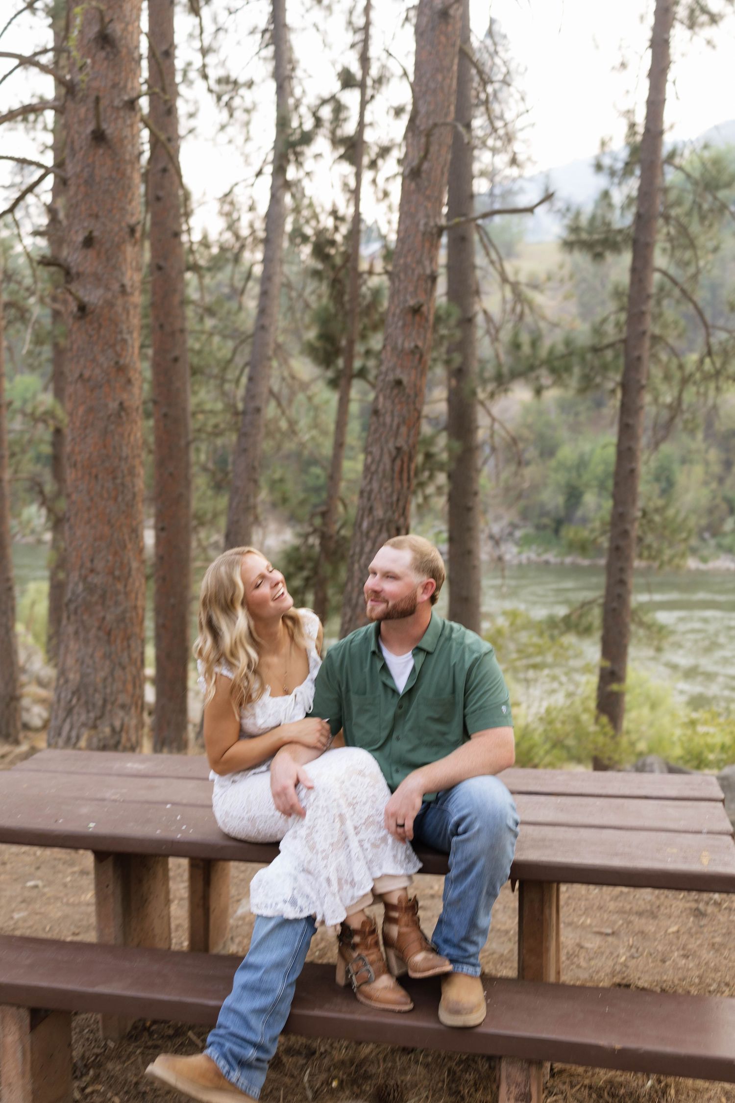 Couple sitting on a wooden picnic table in a pine forest with a river backdrop, woman in white dress and man in green shirt.