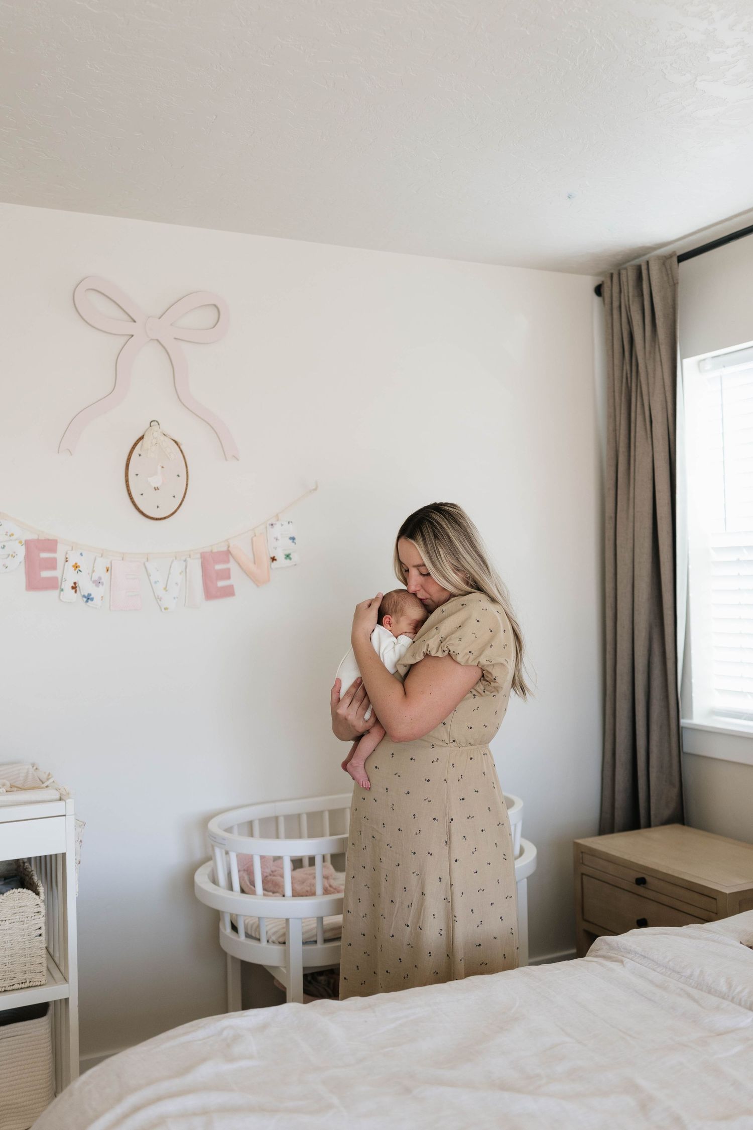 A mother in a beige dress cradles her newborn baby beside a white bassinet in a softly decorated nursery with a pastel banner.