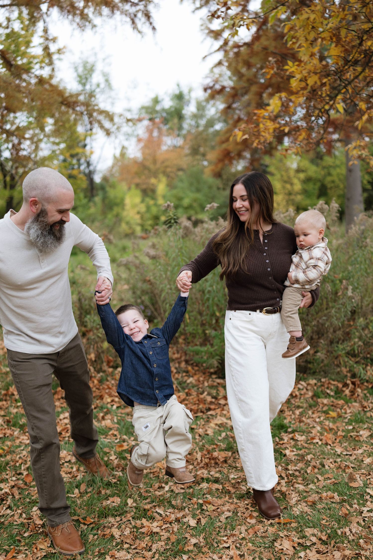 Smiling family of four walks through fall leaves while parents swing a young boy by the hands and mom carries a baby.