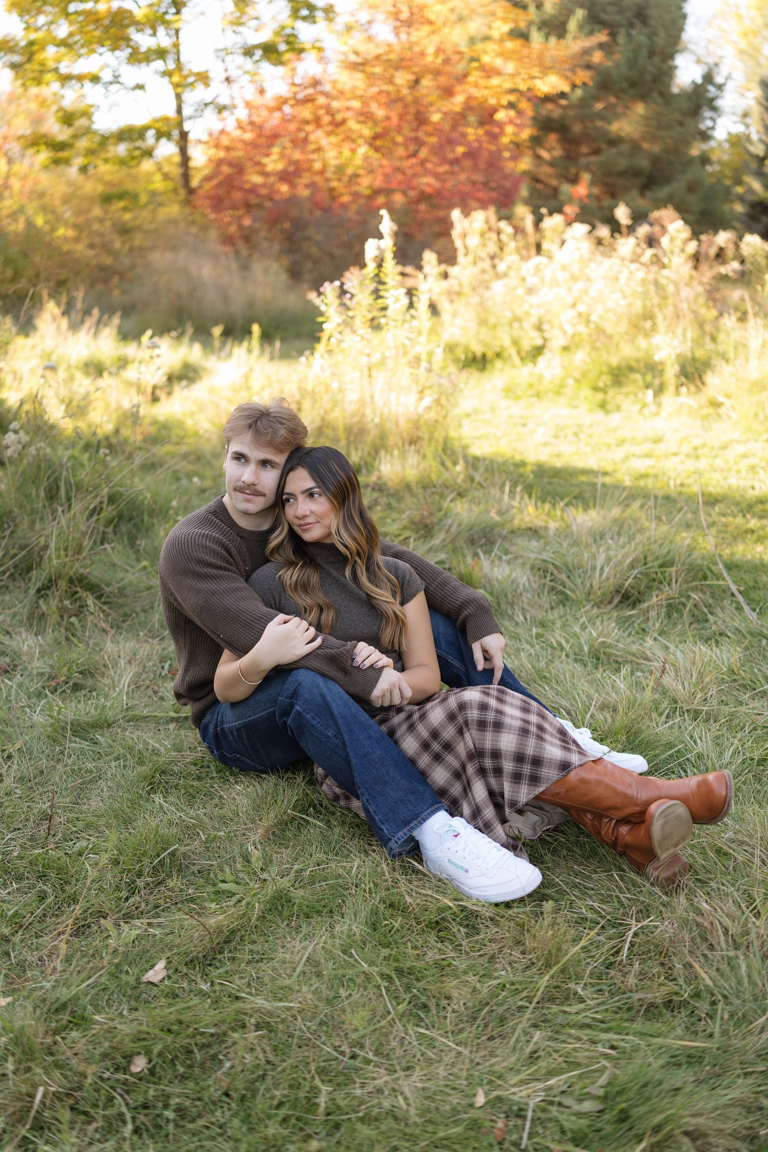 Couple in cozy fall outfits sit close together on a grassy field, with colorful autumn trees glowing behind them.