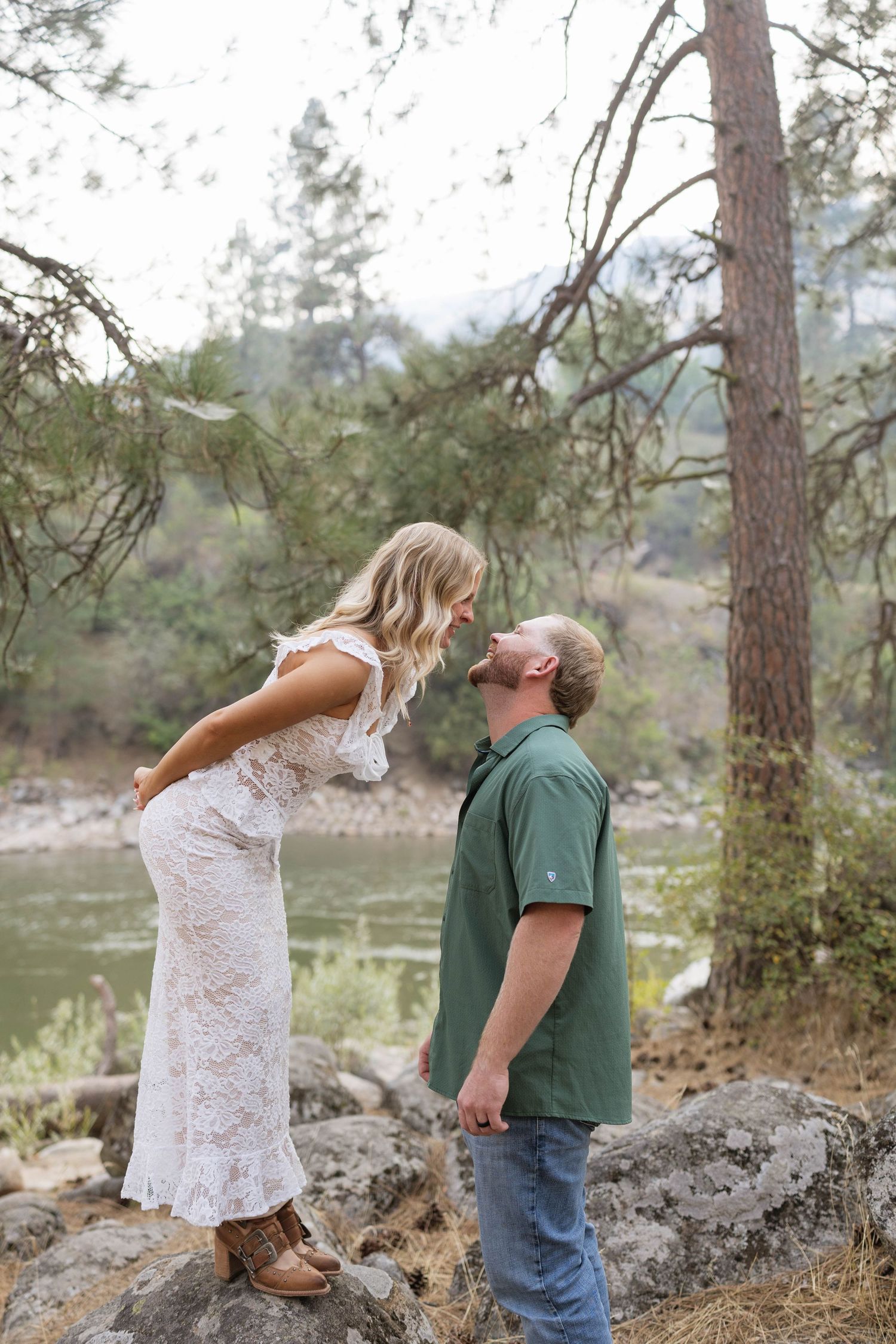 Couple in a lace dress and green shirt lean in for a kiss on a rocky riverbank surrounded by pine trees.