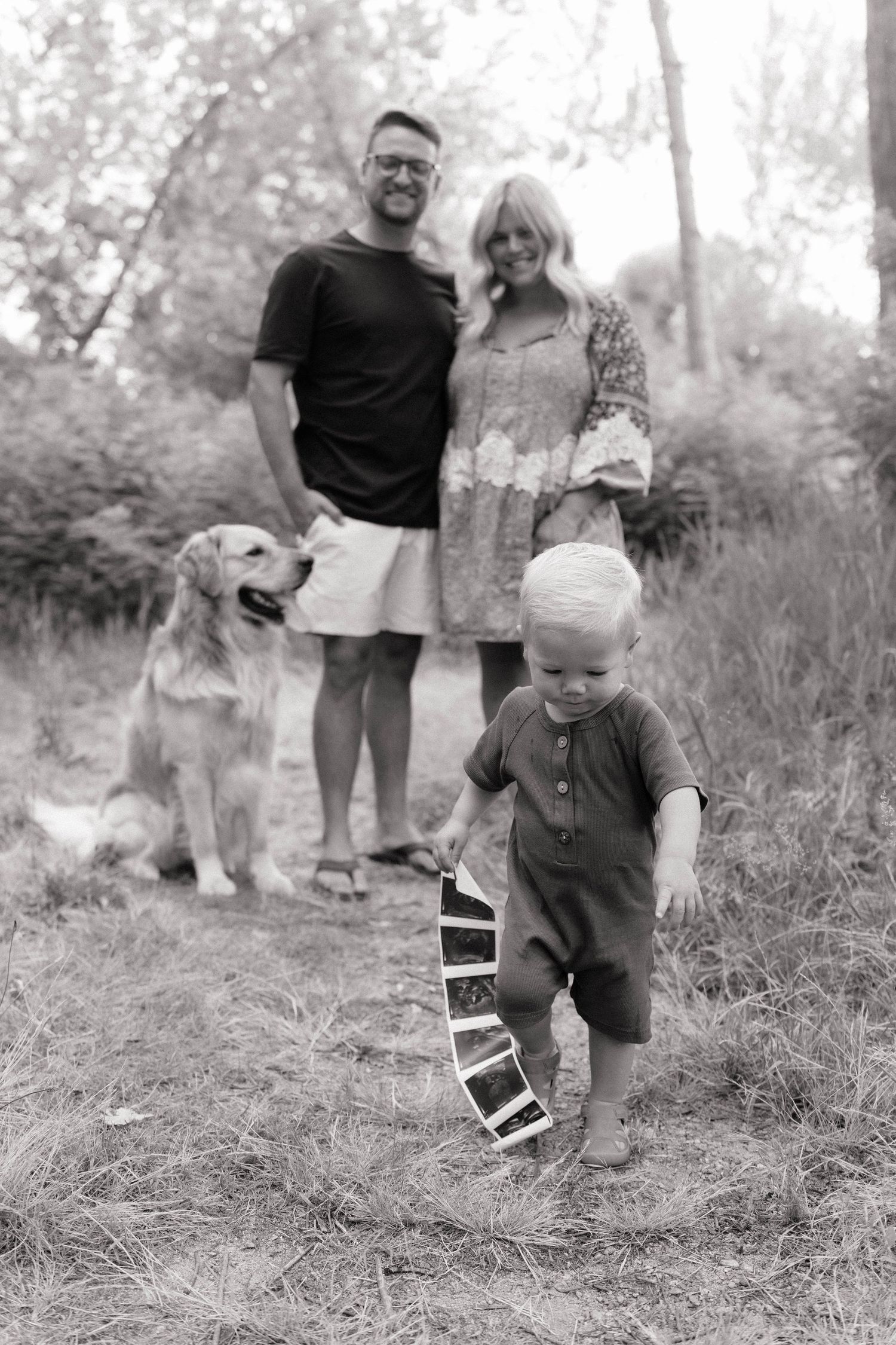 Black and white family photo with parents, golden retriever, and toddler holding a strip of photos outdoors.