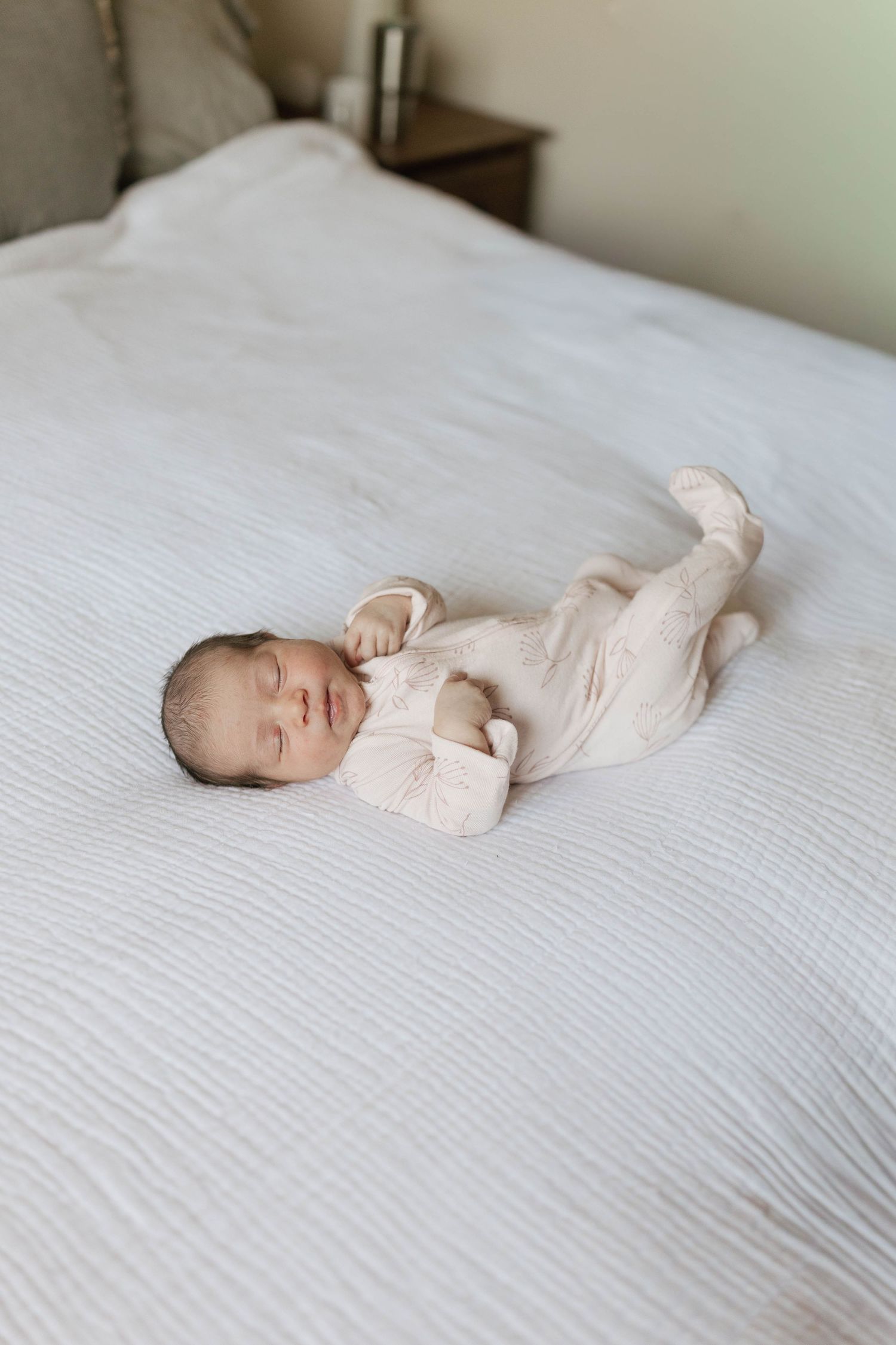 A newborn baby wearing a light beige onesie lies peacefully on white bedding in a series of gentle poses.