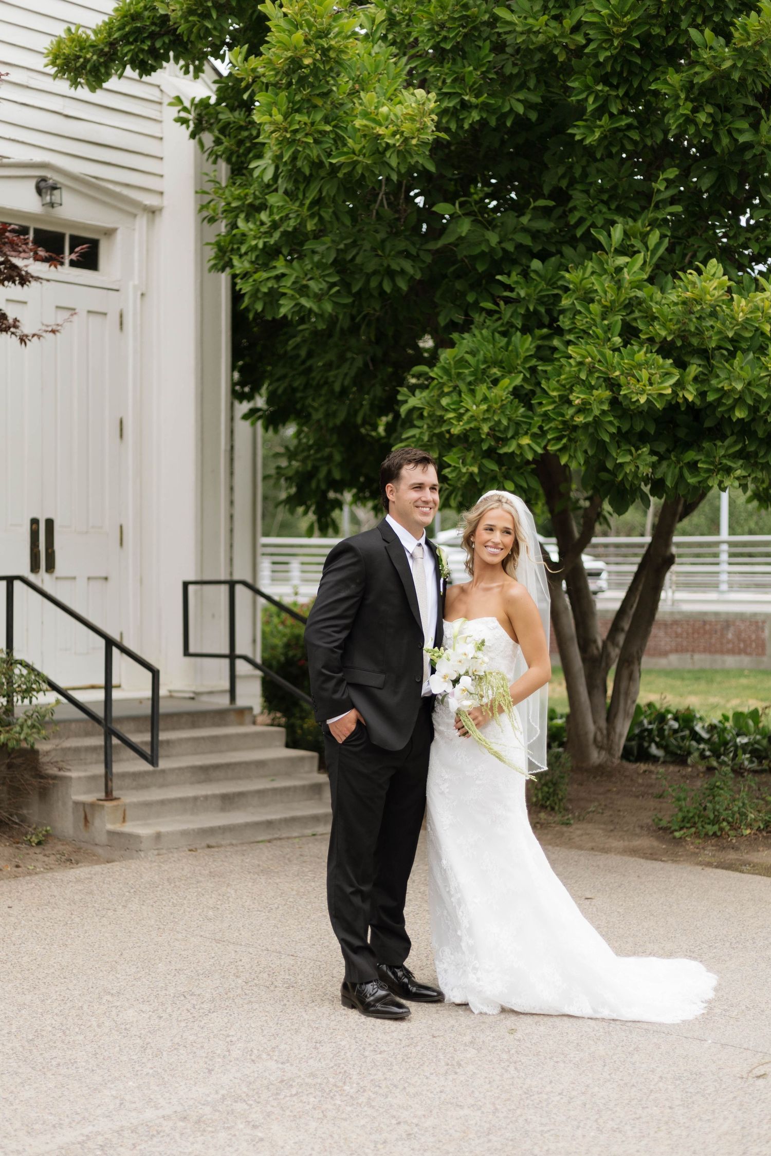 Bride and groom posing together outside a white chapel, bride holding white bouquet in elegant strapless gown.