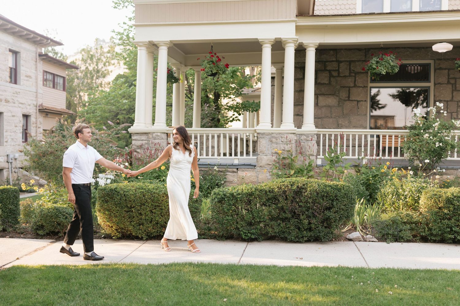 A couple holds hands and walks along a sidewalk in front of a charming stone house with white columns.