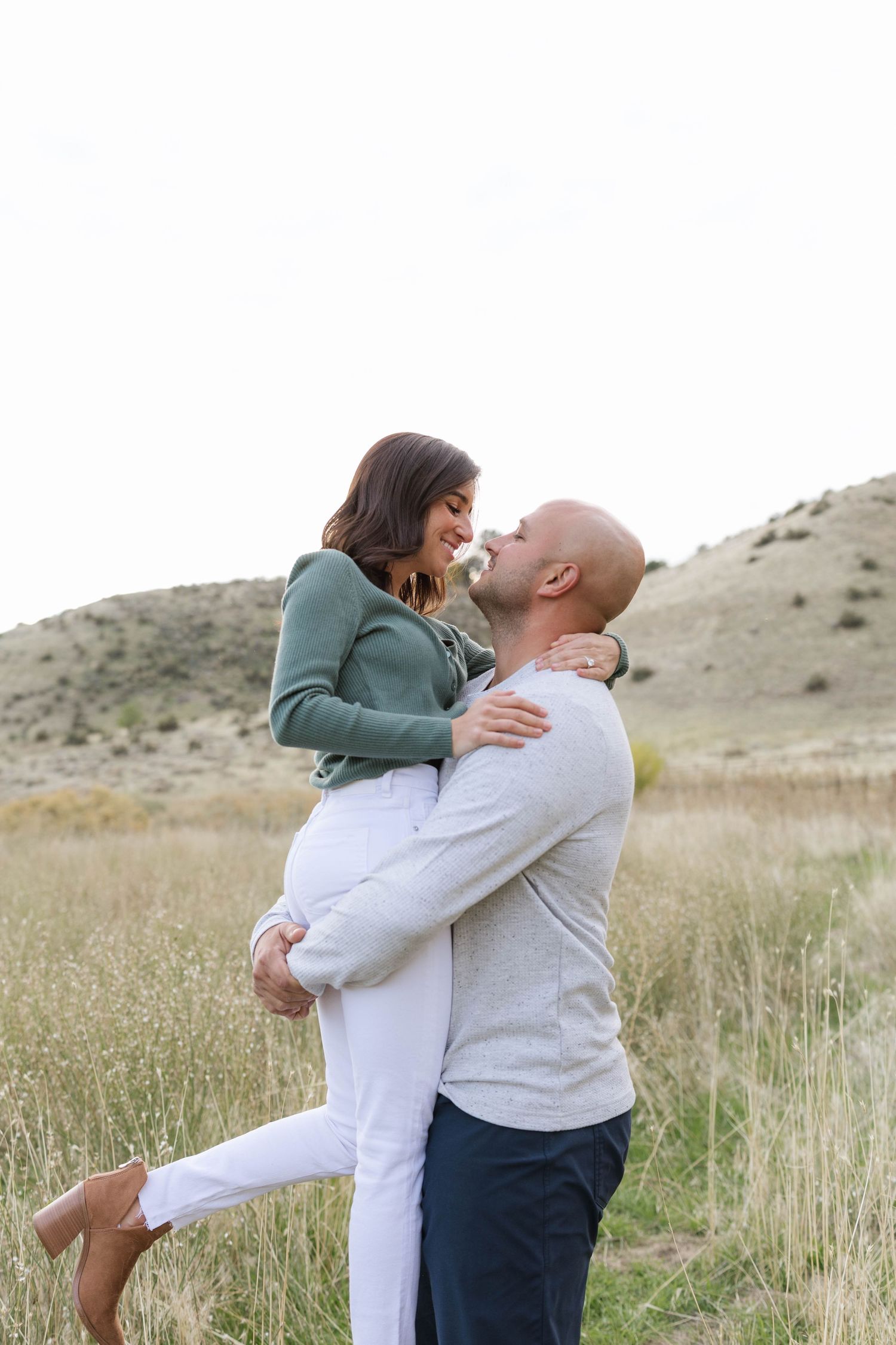 A couple embraces lovingly in a field of tall grass with rolling hills in the background.