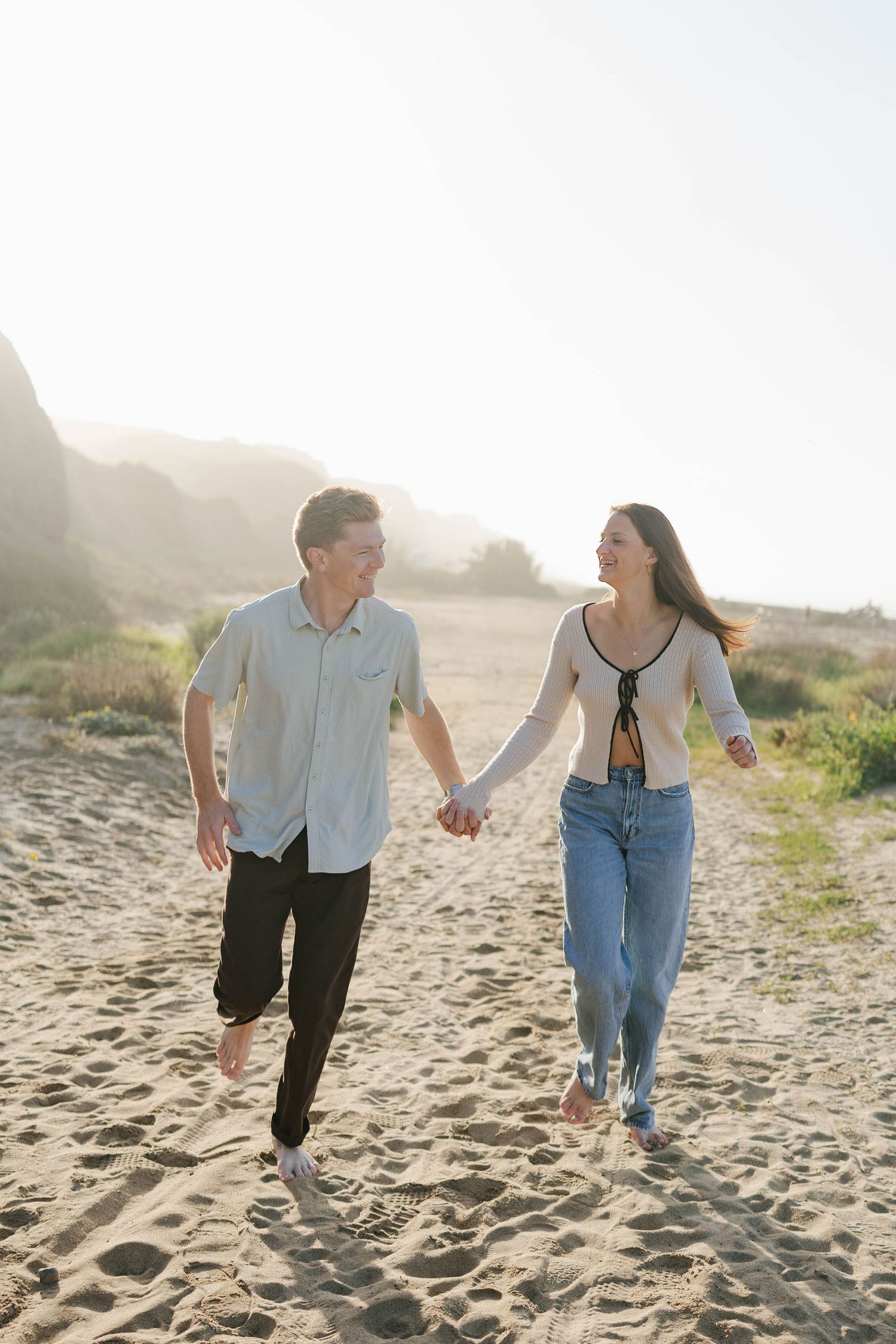 A couple runs playfully along a sandy beach at sunset while holding hands and smiling at each other.