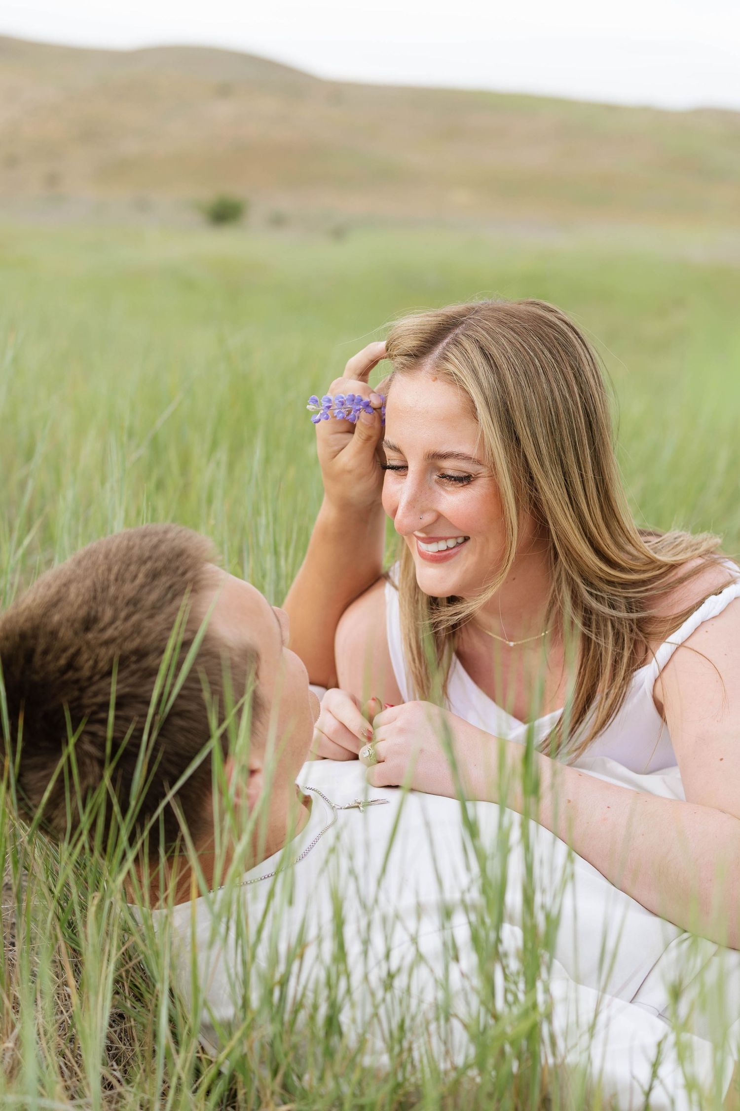 Smiling blonde woman holding purple wildflowers while sitting in tall green grass with a companion.