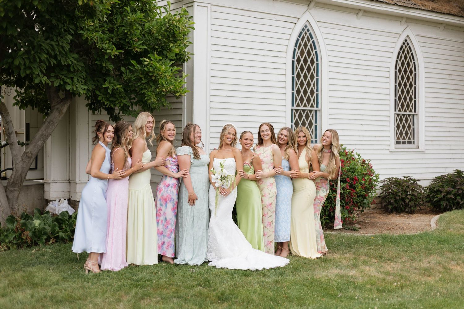 Bride poses with bridesmaids in pastel dresses outside a white chapel on a sunny wedding day.