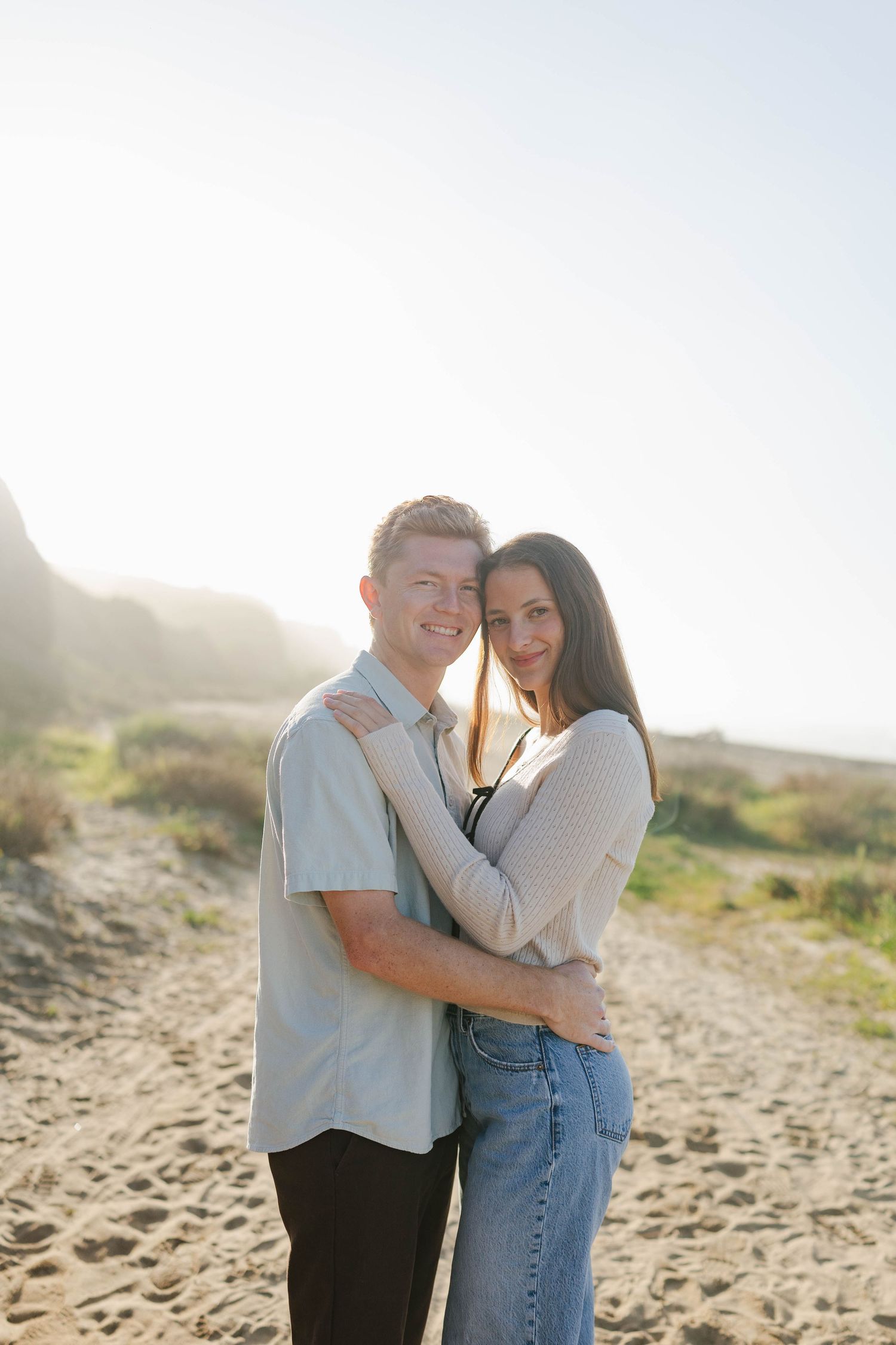 A couple embraces on a sunny beach with soft lighting and sandy dunes in the background.
