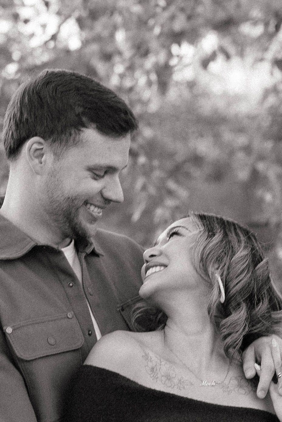 Black and white portrait of a smiling couple gazing at each other outdoors, the woman leaning back against the man with trees behind them.