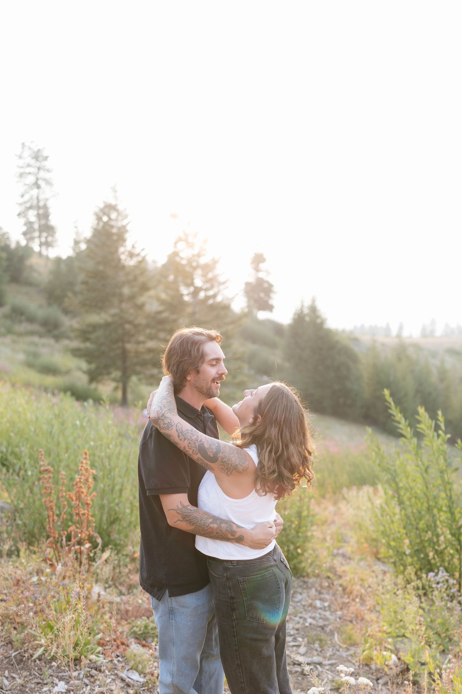 Smiling couple embracing in a sunlit mountain meadow with evergreen trees, capturing a warm, romantic engagement photo moment.