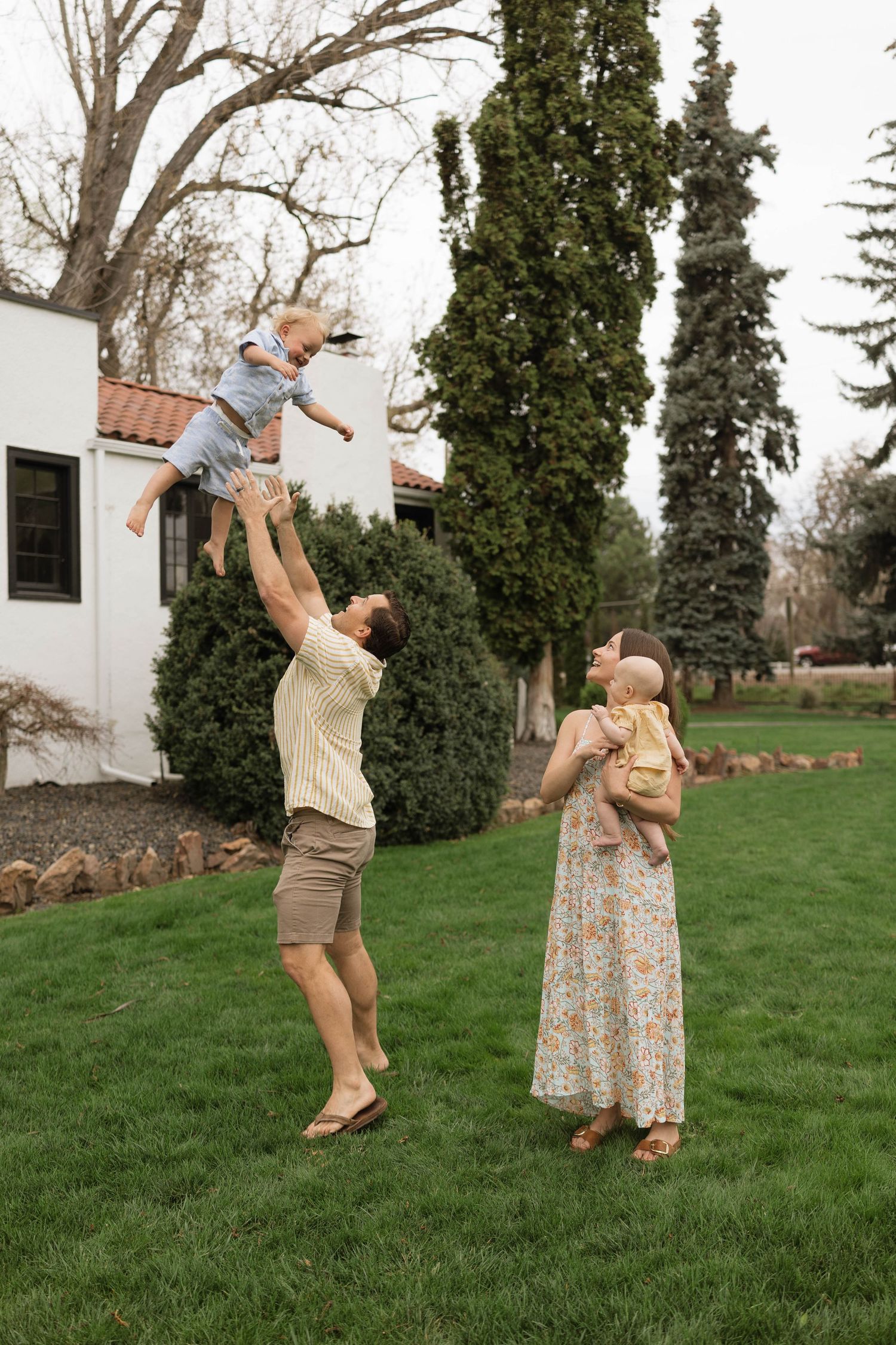 A family plays together on a green lawn outside their home with tall evergreen trees in the background.