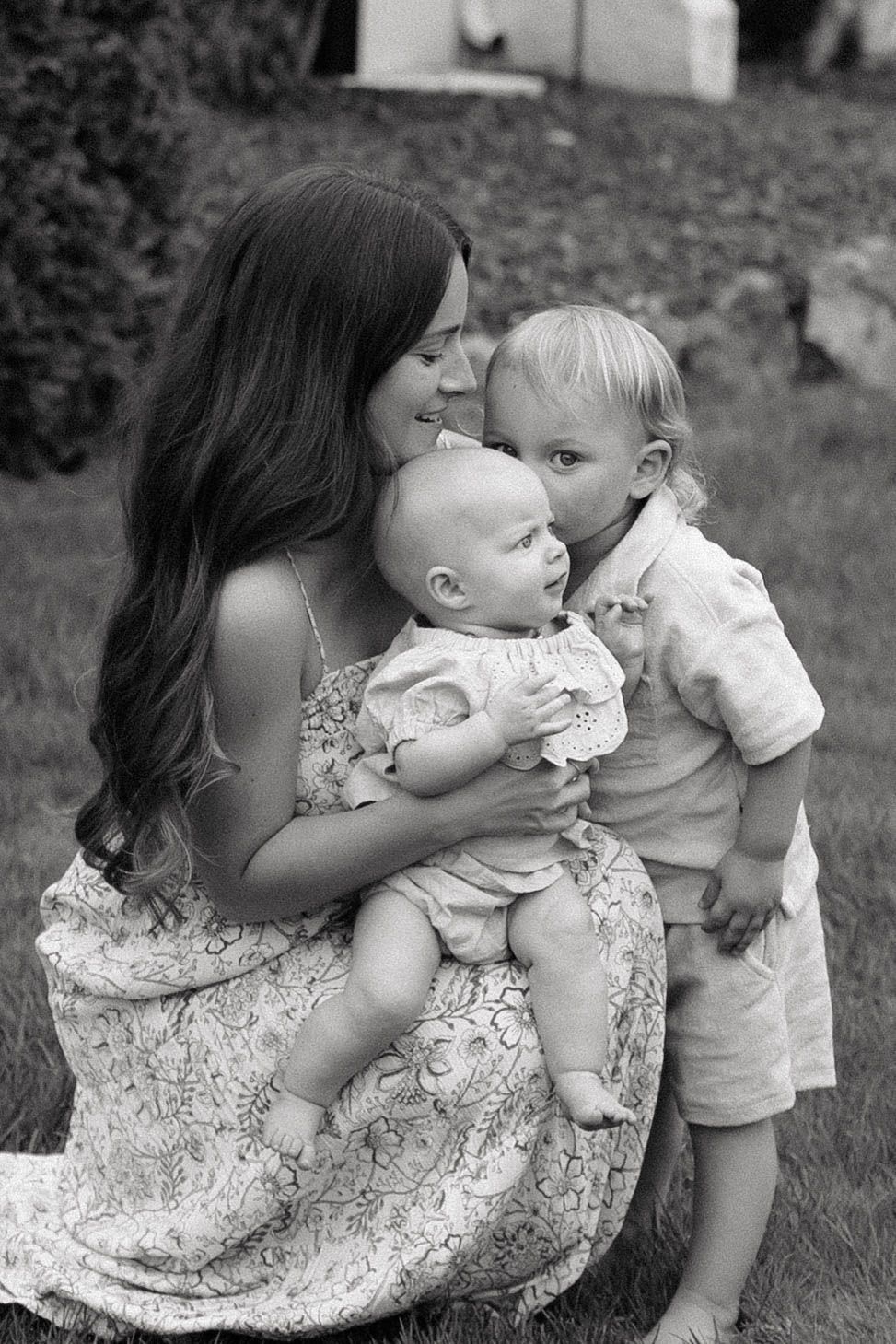 Black and white photo of a young mother sitting on grass, tenderly holding an infant while a toddler leans in close.