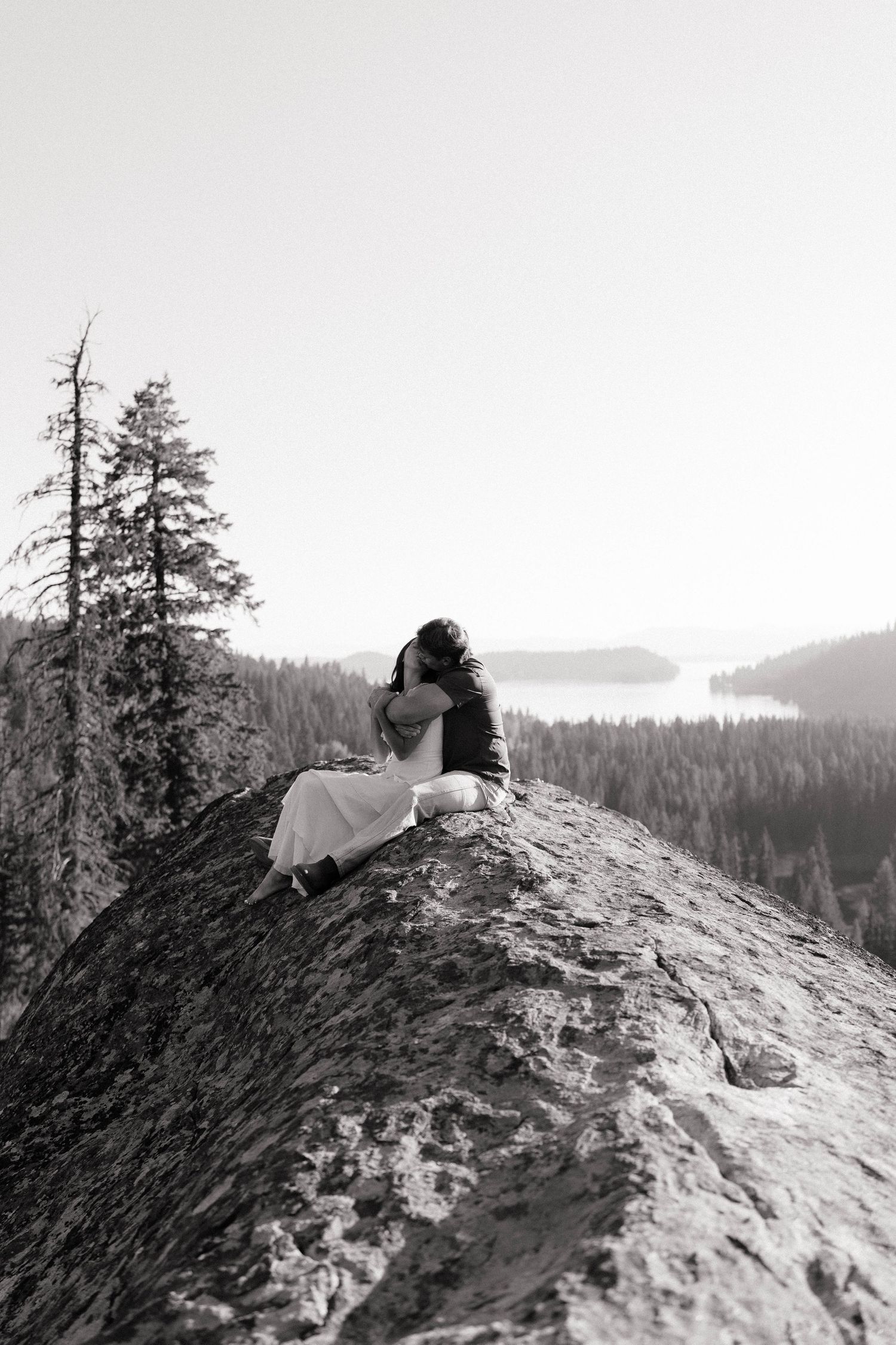 A person lies on a rocky mountain peak overlooking a vast forested wilderness landscape in black and white.