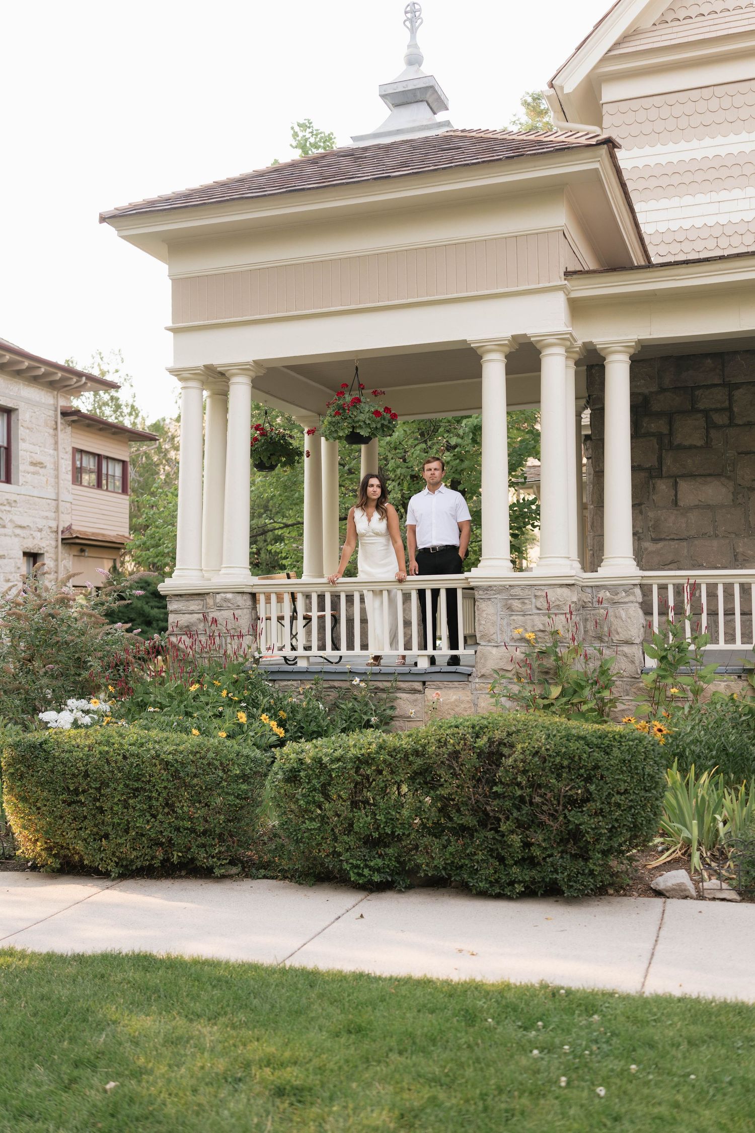 Couple standing on elegant Victorian home porch with white columns, surrounded by lush gardens and colorful flowers.