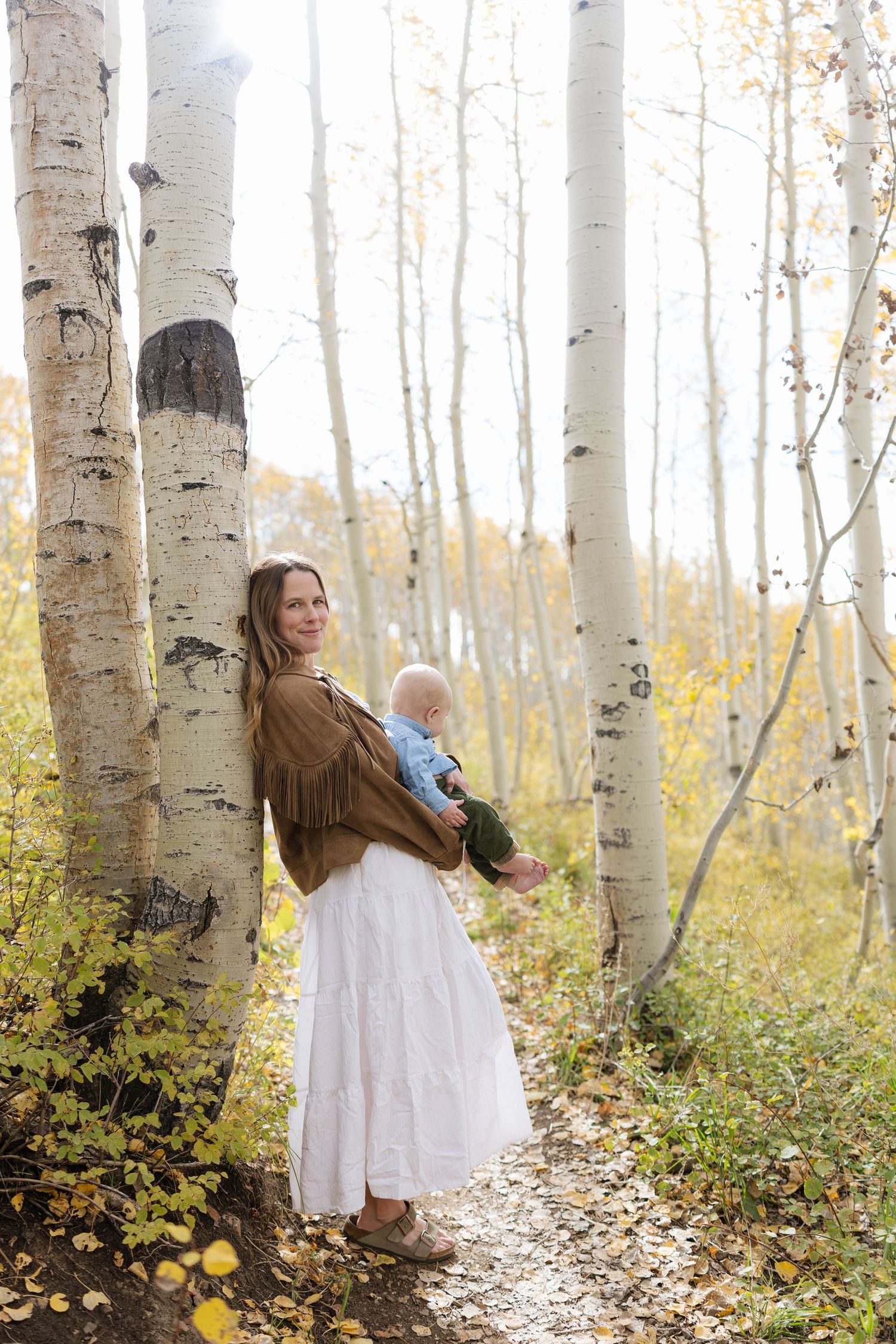 A mother in a white skirt and fringe jacket holds her baby among white aspen trees with golden fall foliage.