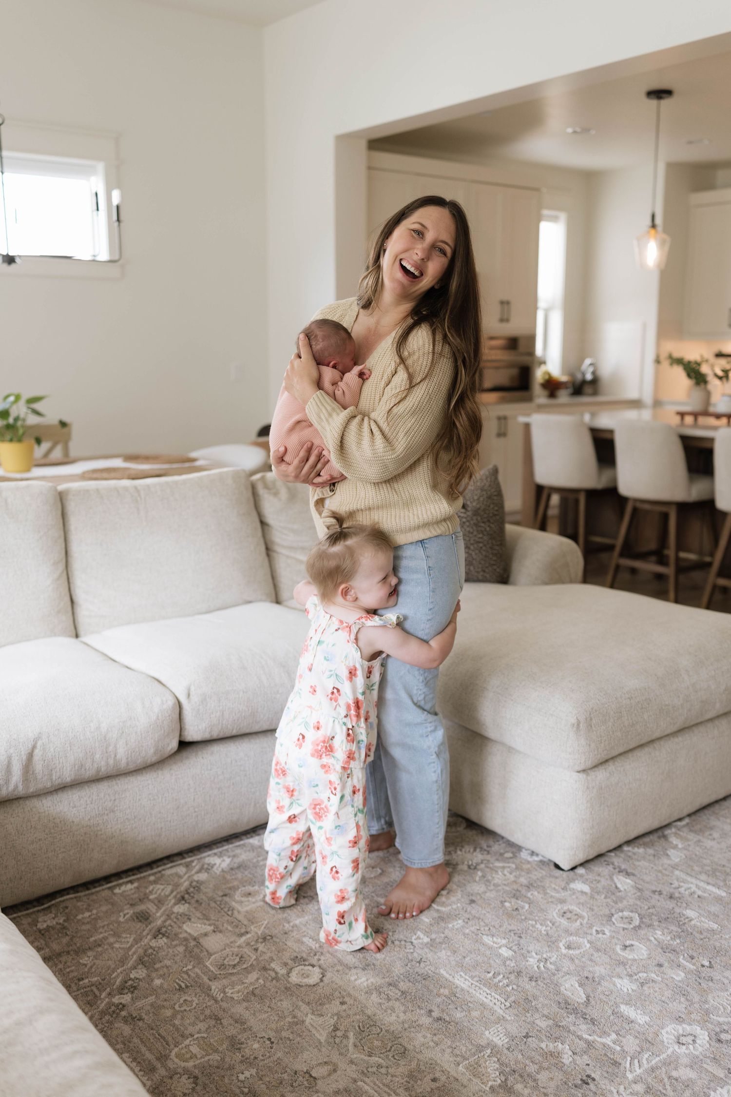 Smiling woman holding newborn baby while toddler in floral pajamas hugs her legs in a bright modern living room.