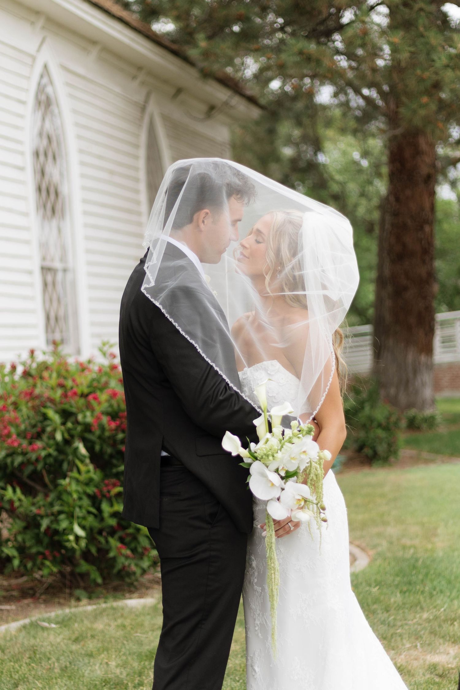 Bride and groom kissing under a white veil outdoors, bride holding white floral bouquet near a white church.