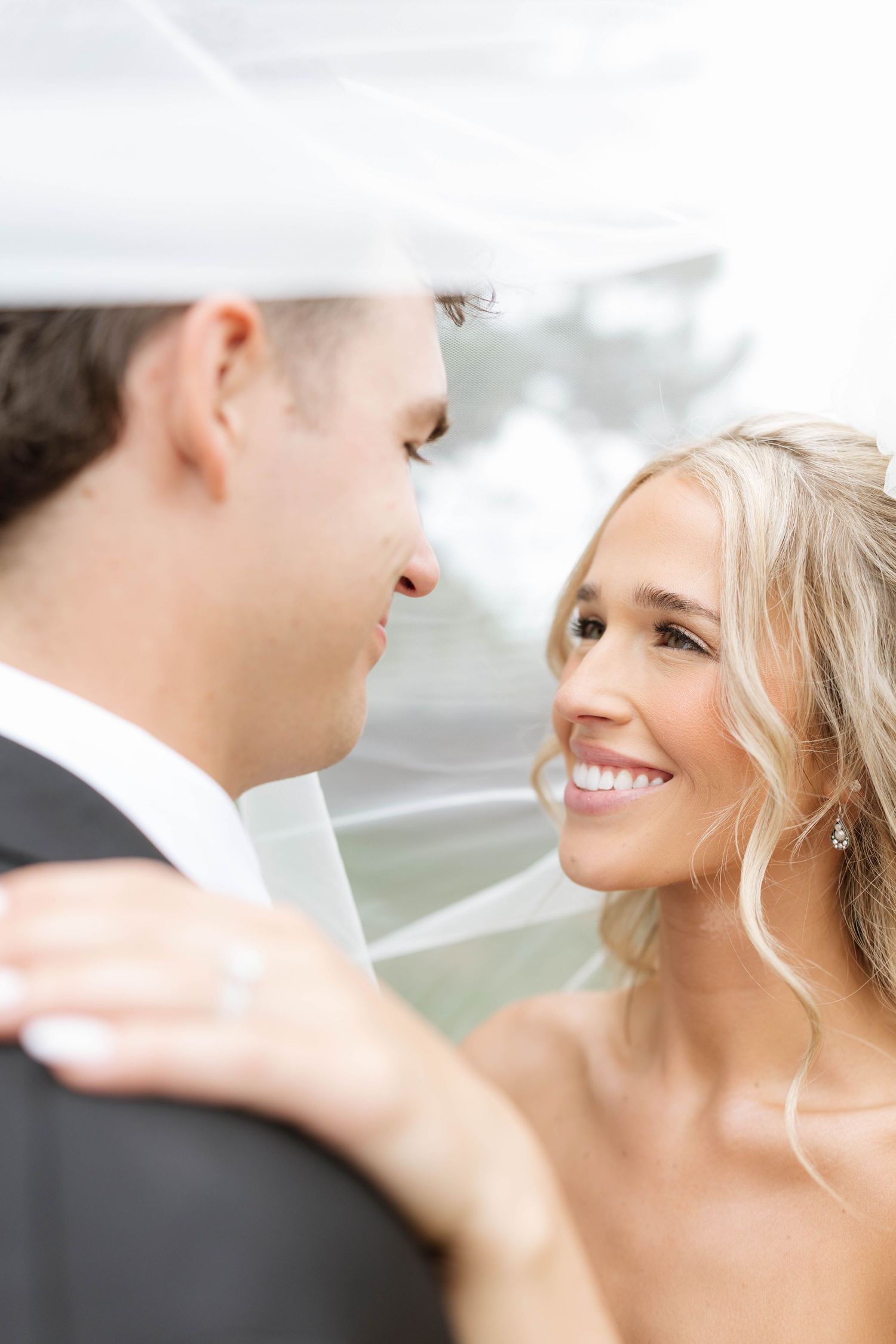Bride smiling at groom under wedding veil during romantic outdoor portrait session.