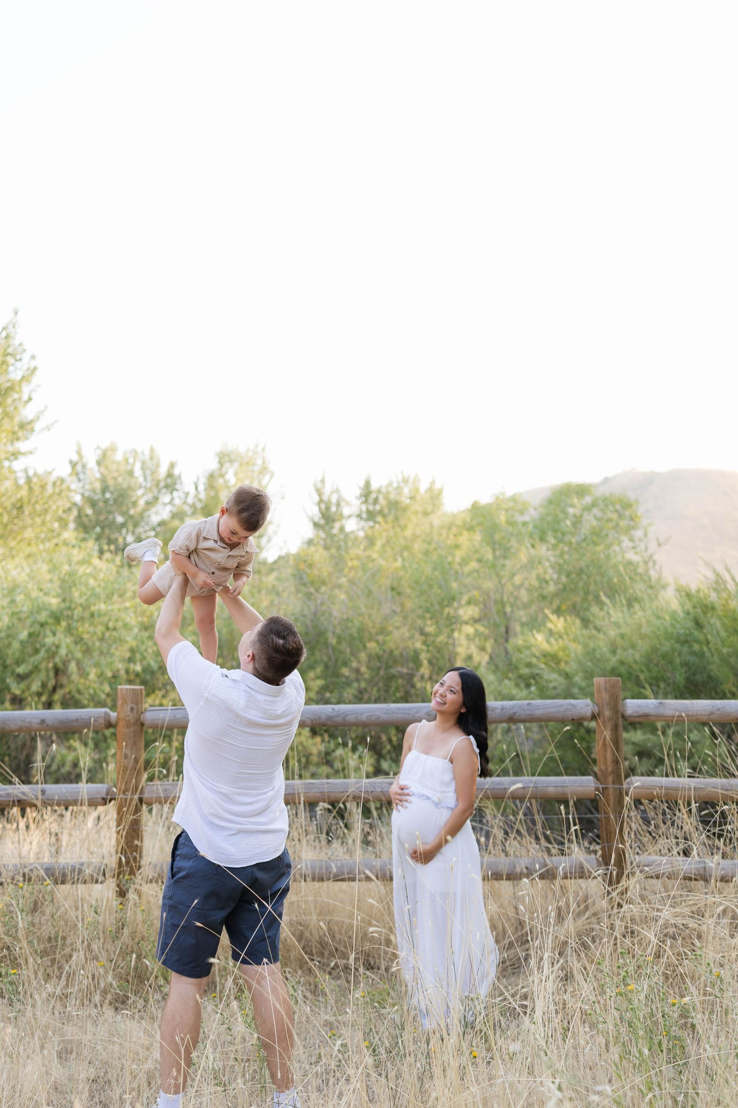 A father lifts his baby in the air while a pregnant mother in a white dress smiles outdoors near a wooden fence.