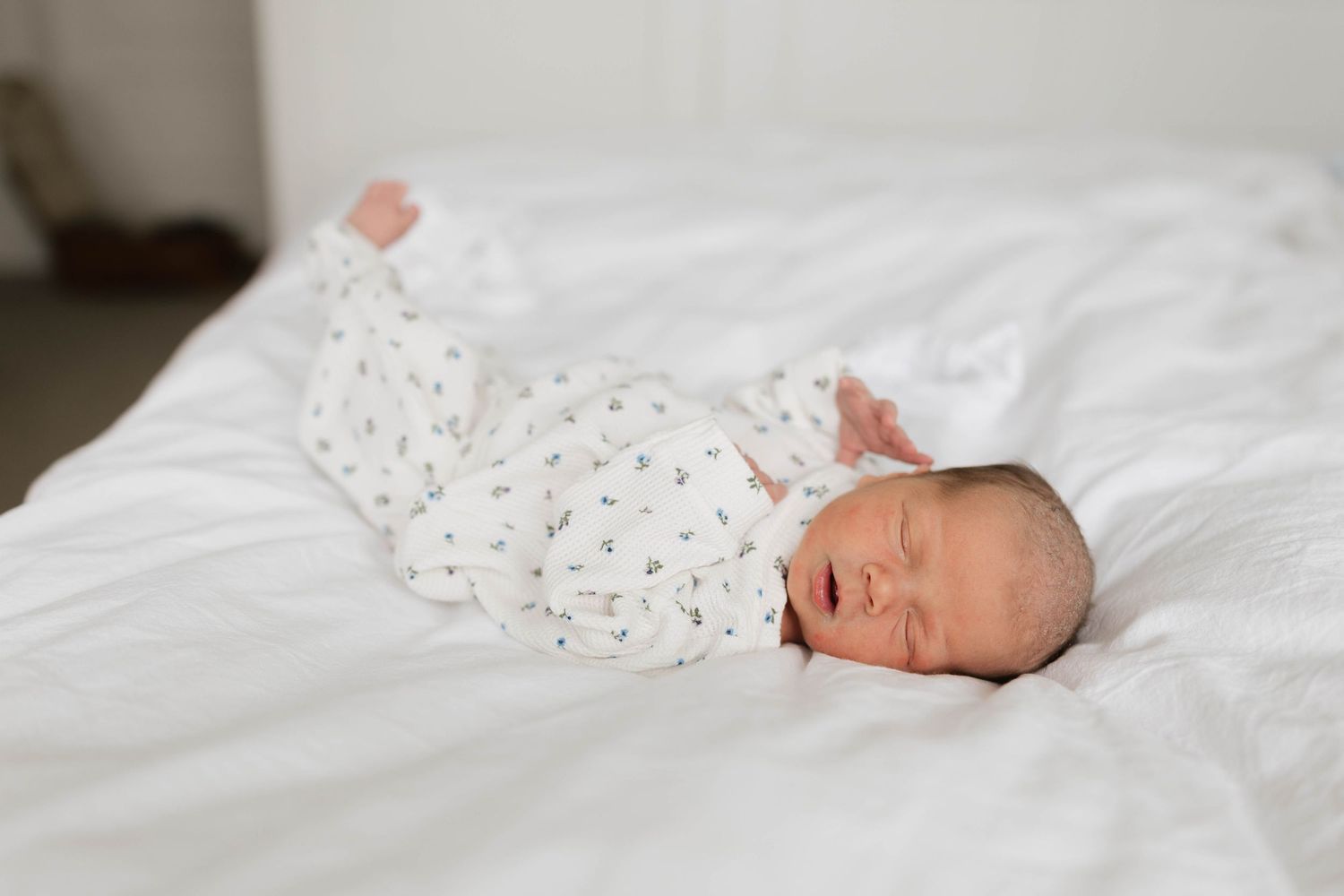 Newborn baby sleeping peacefully on white bedding wearing a white floral onesie with arms raised.
