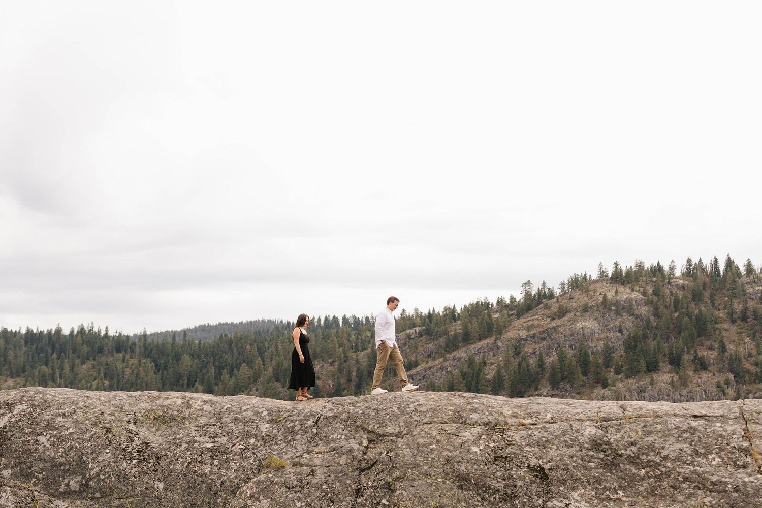 Couple walking toward each other along a rocky ridge overlooking a forested valley under a soft overcast sky.