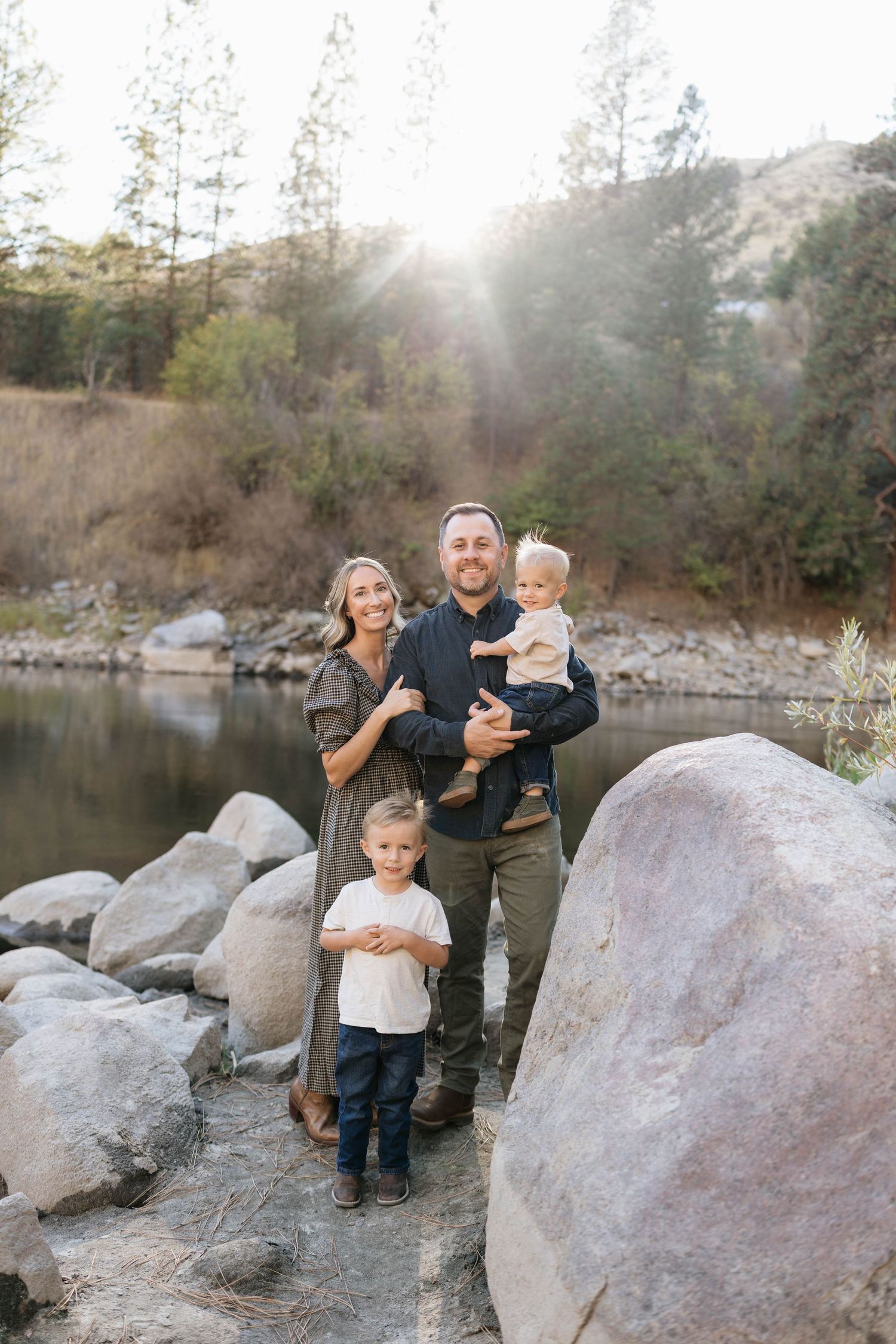 Smiling family of four with two young boys poses among large boulders beside a calm river, with trees and golden sunlight behind them.