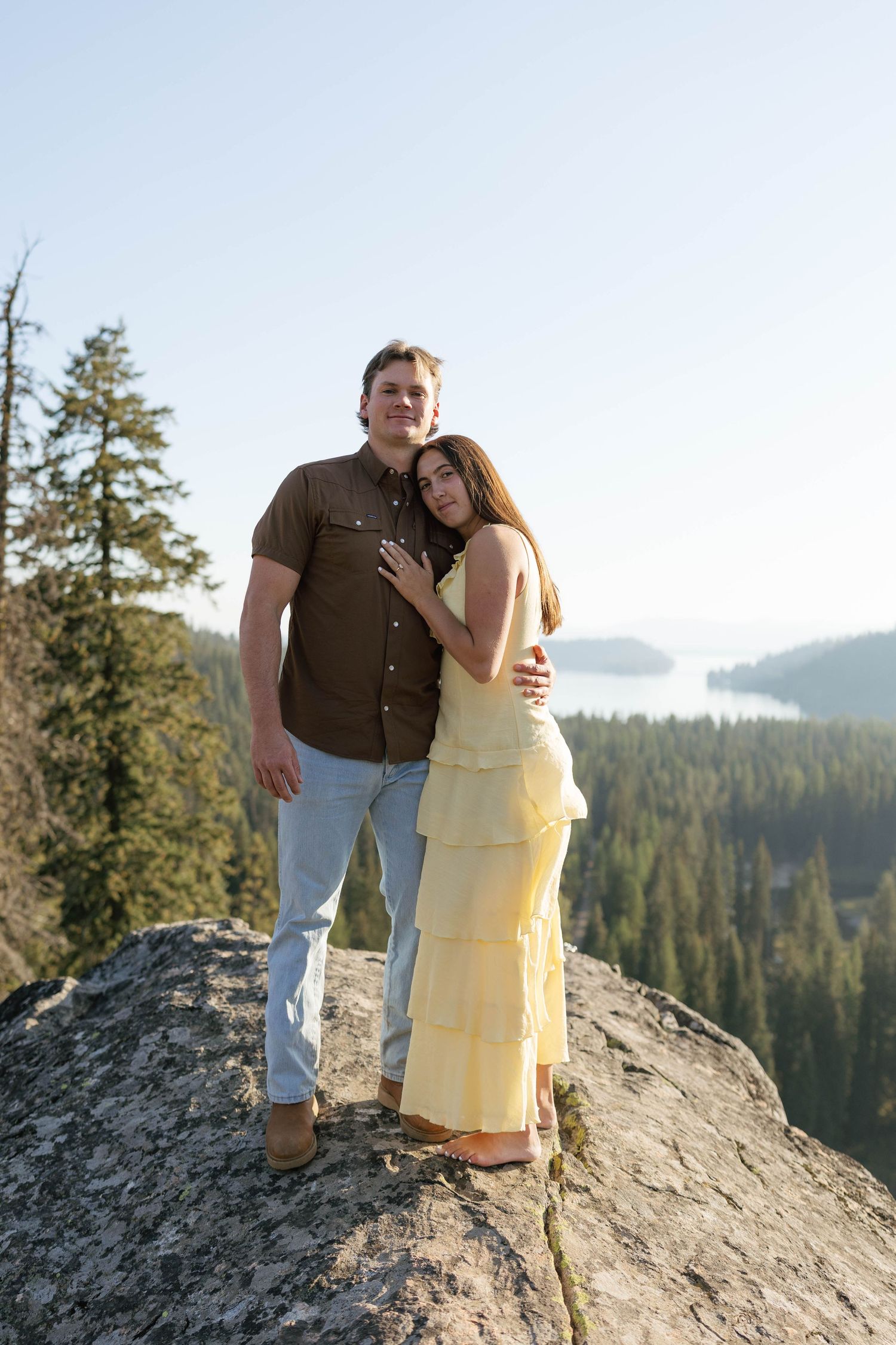 Couple embraces while standing on a rocky mountain peak overlooking a scenic forest landscape on a sunny day.