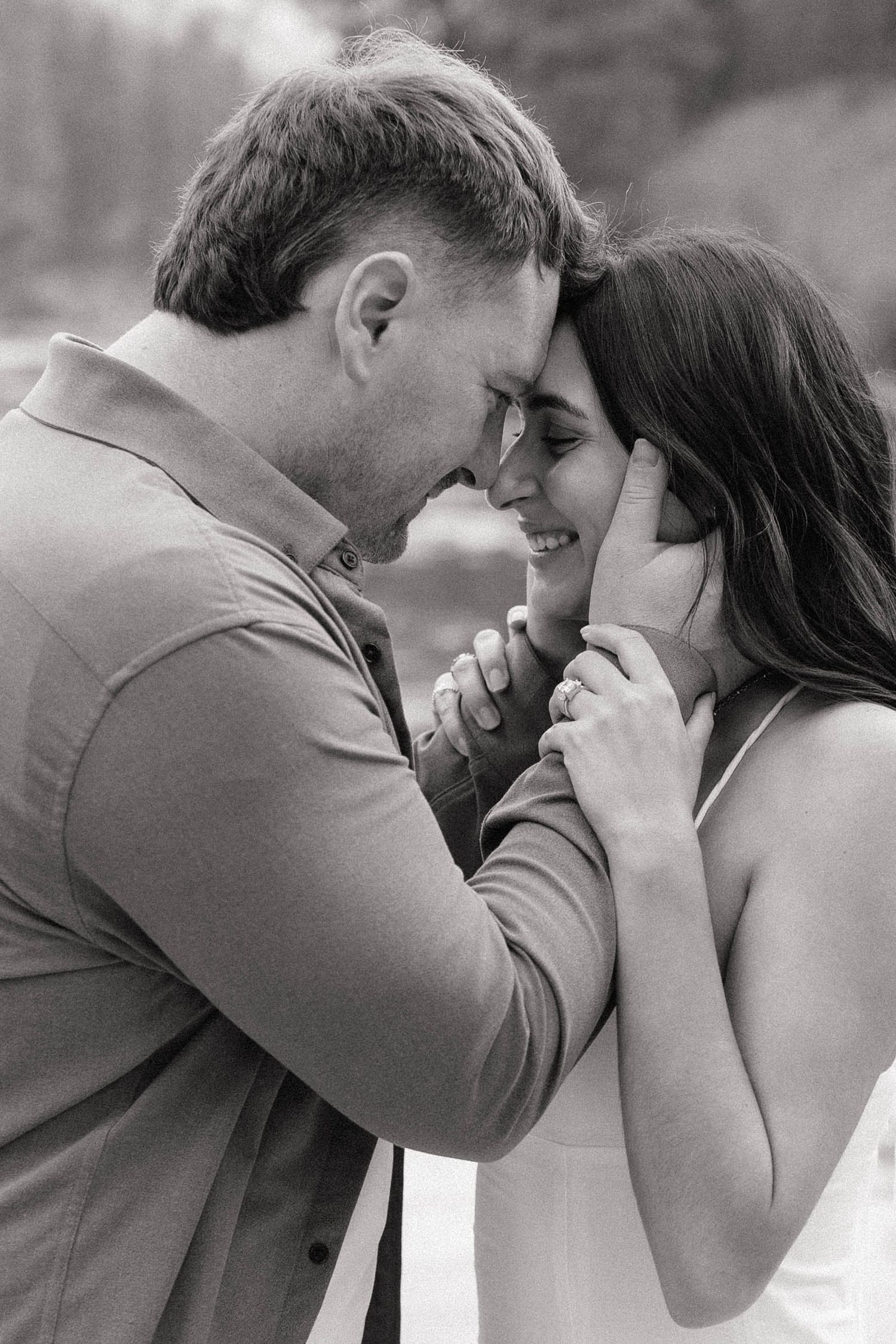 Black and white photo of a romantic couple touching foreheads and holding hands, sharing an intimate moment outdoors.