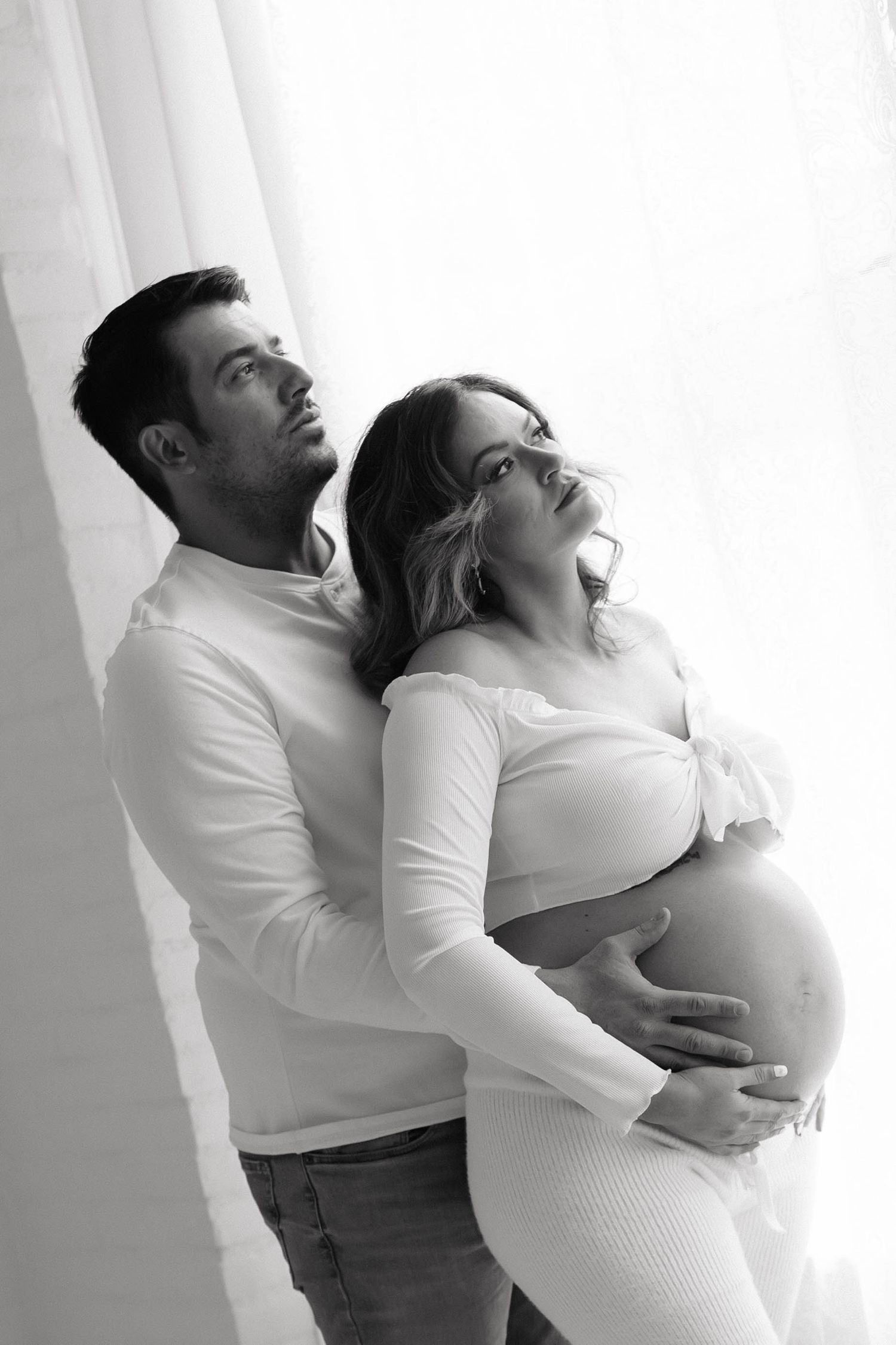 Black and white maternity photo of a couple, man embracing pregnant woman from behind near a bright window.