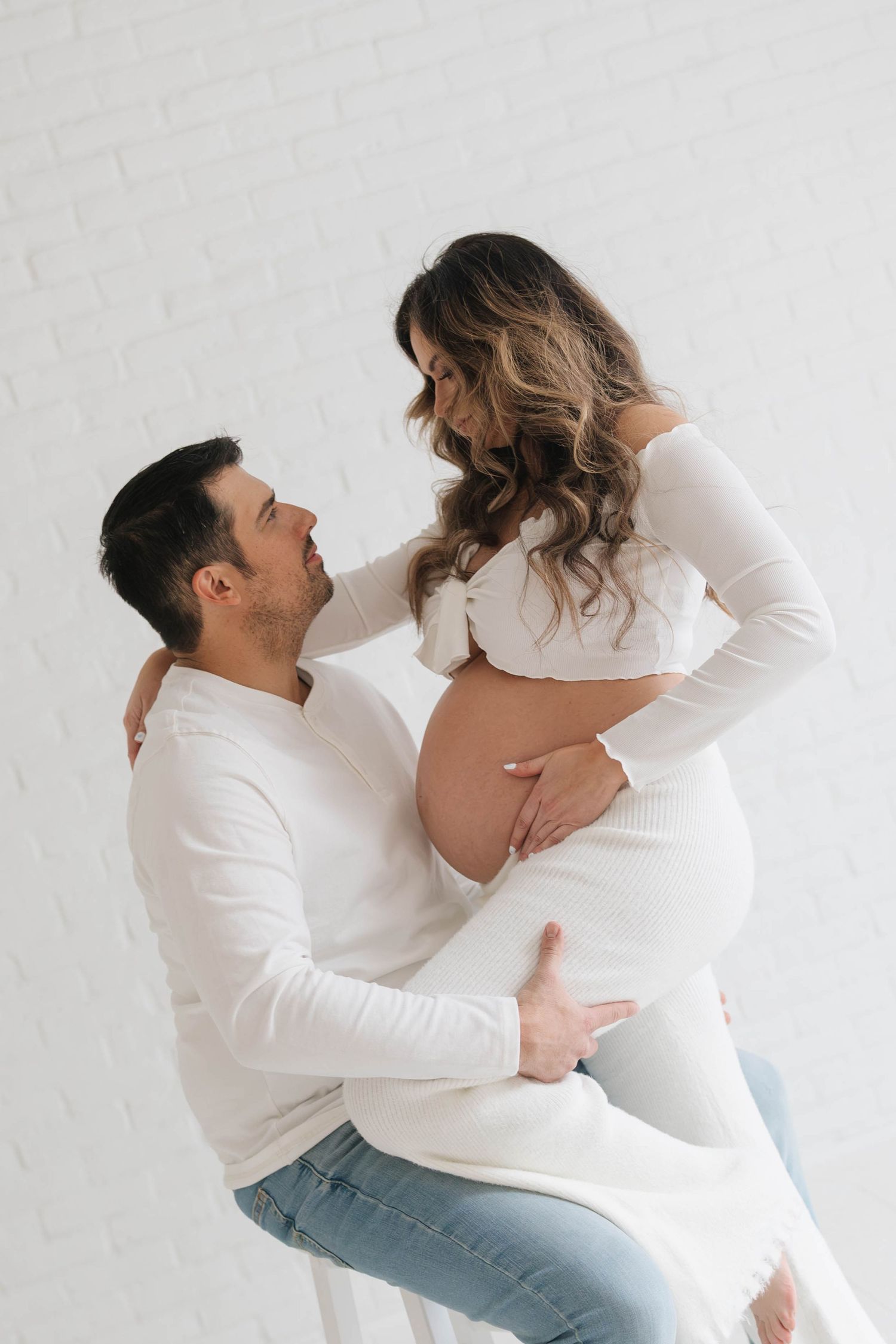 Intimate maternity photo of couple in white tops embracing against white wall.