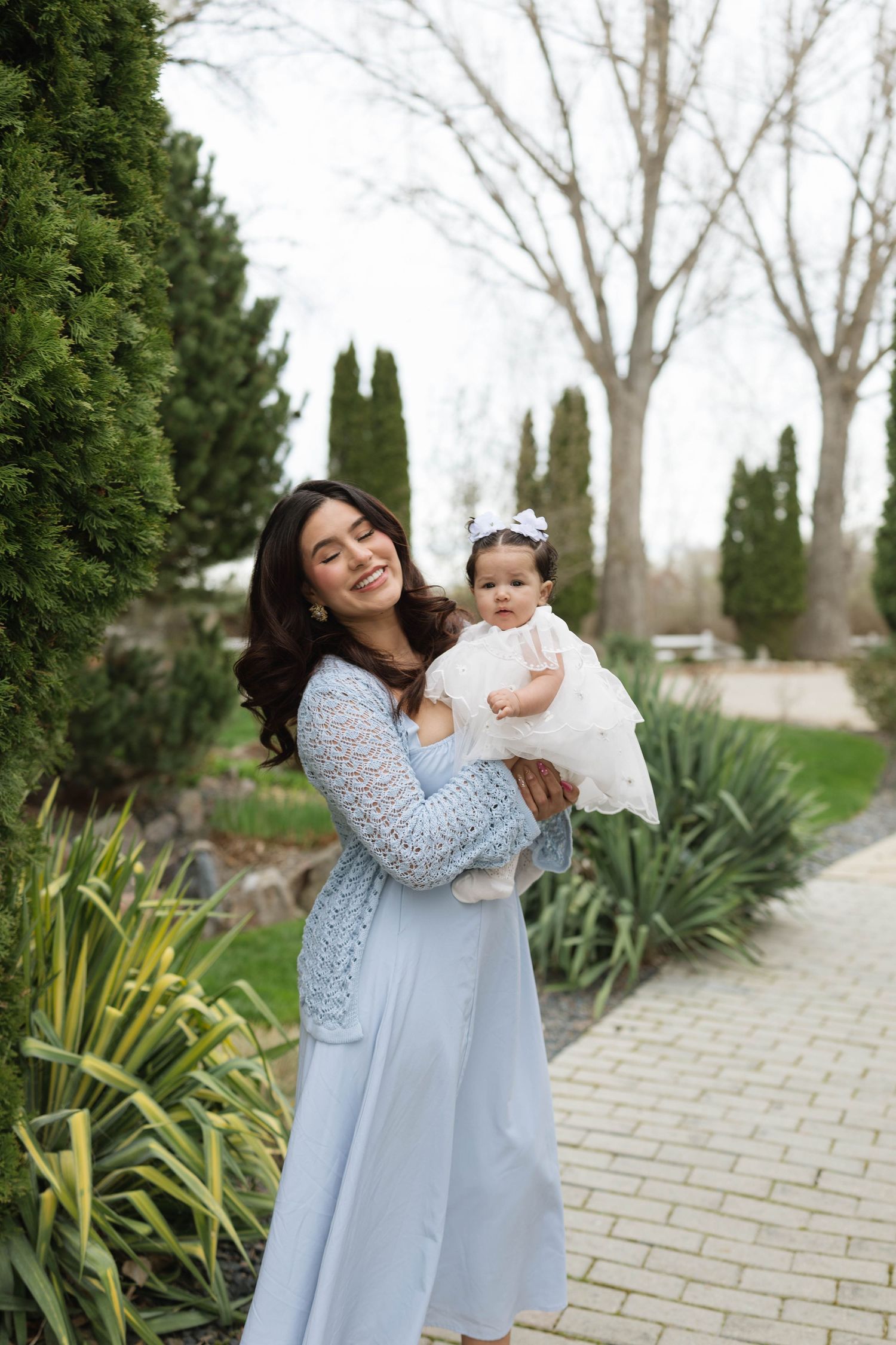 Smiling mother in blue dress holds baby girl in white dress outdoors in a lush green garden setting.
