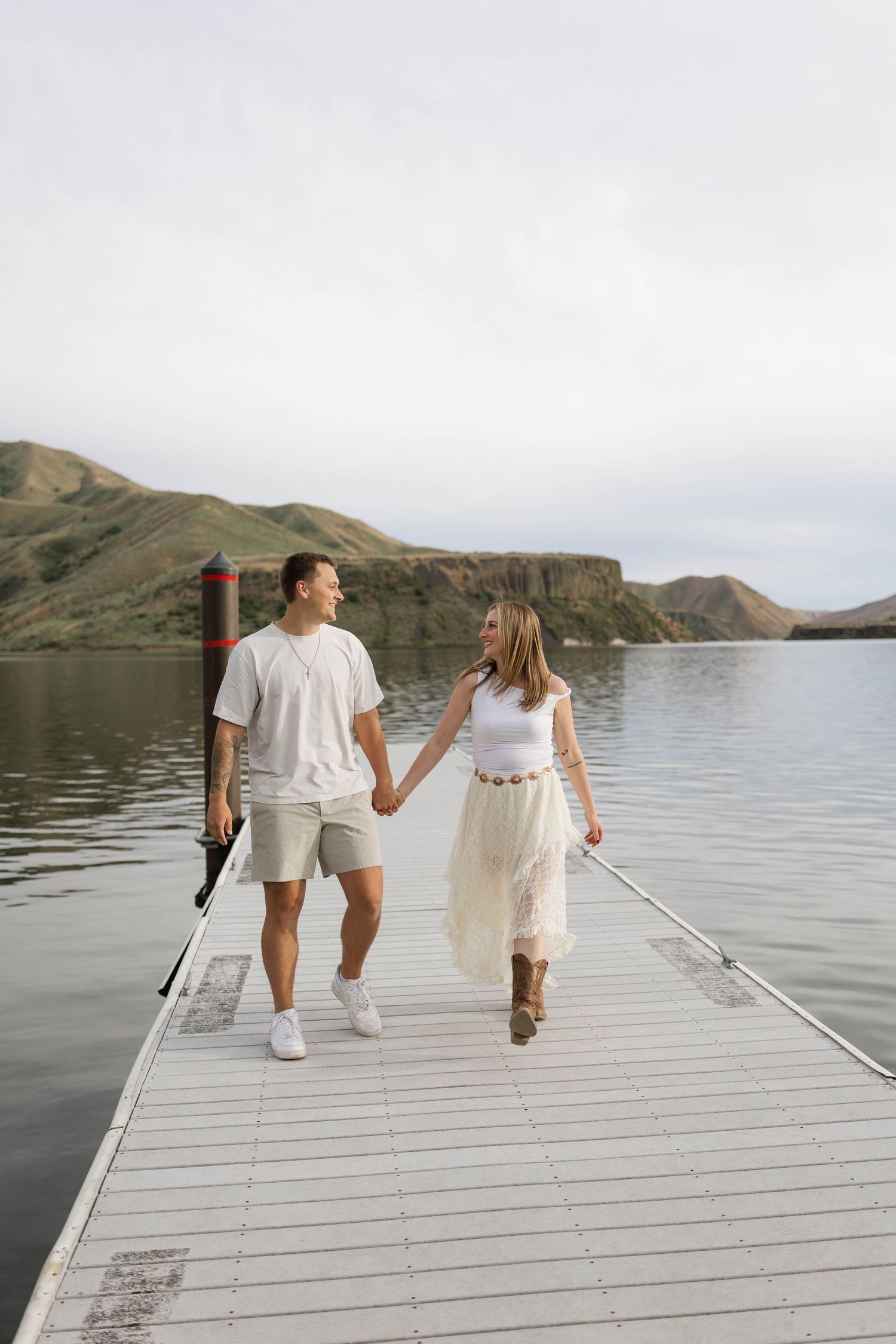 Couple holding hands walking on a dock by a calm lake with scenic hills in the background.