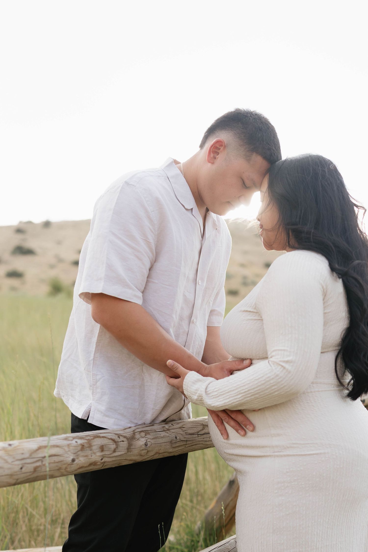 Couple in white outfits sharing a tender moment outdoors, woman in maternity dress holding baby bump near wooden fence.