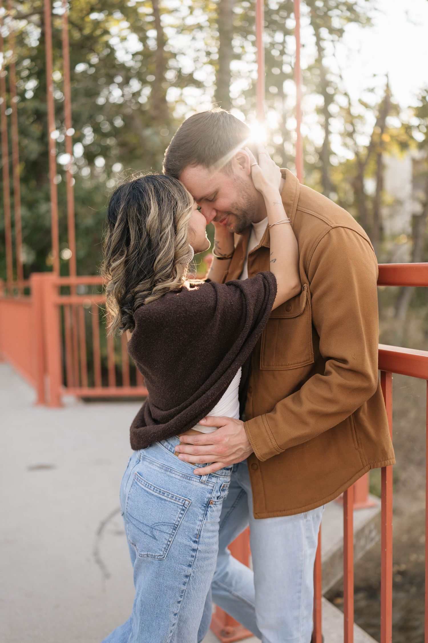 Couple in an affectionate embrace on a red suspension bridge, foreheads touching as warm sunlight filters through the trees.