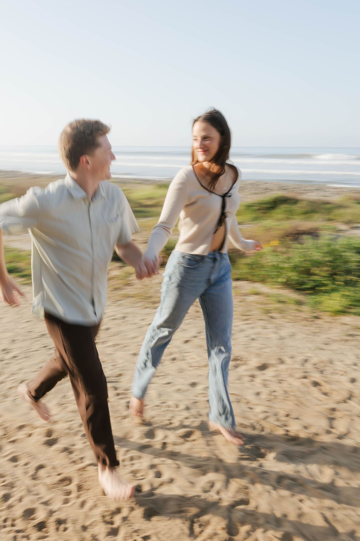 A couple playfully runs and chases each other on a sandy beach with ocean waves visible in the background.