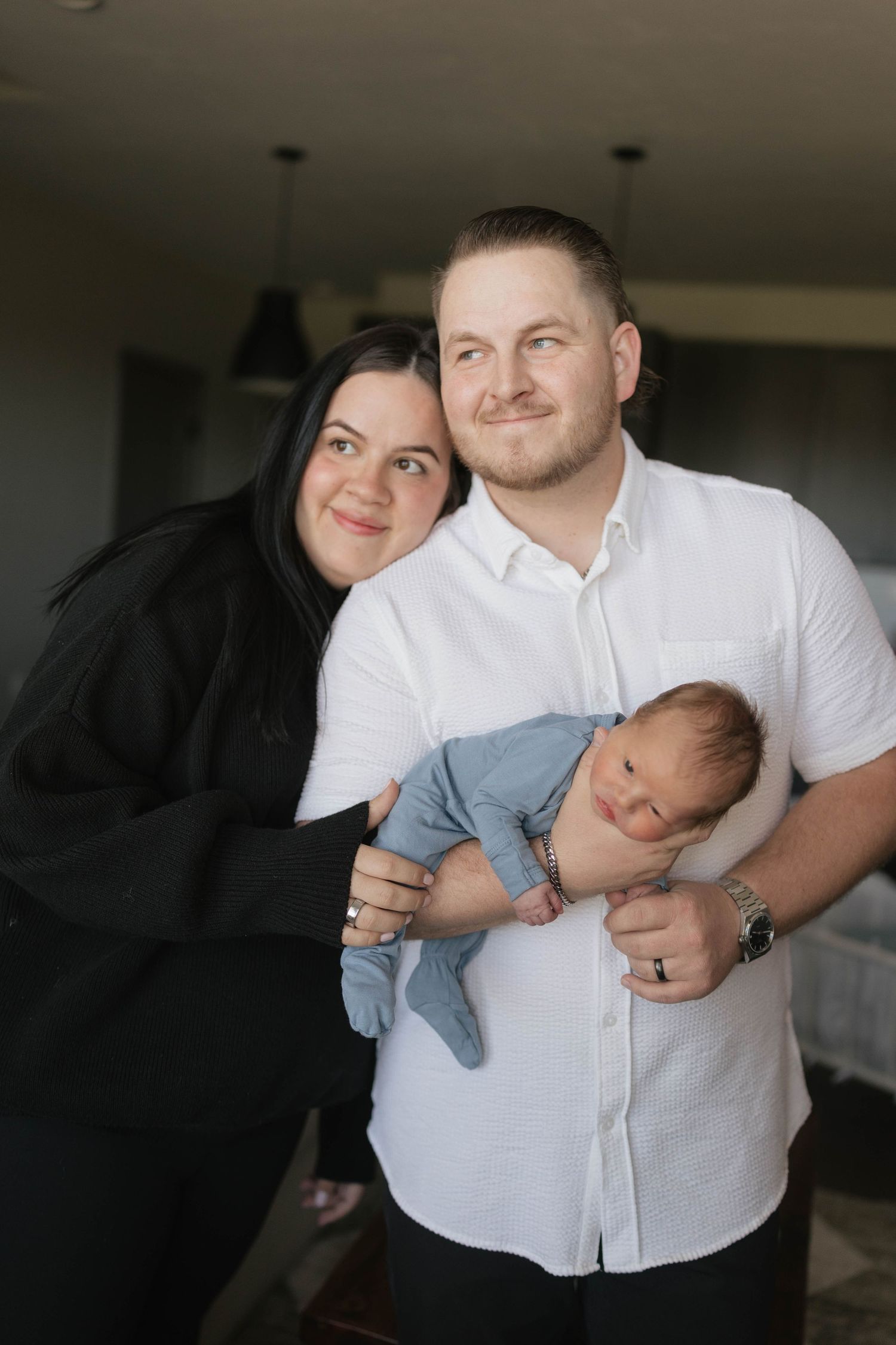 A couple holds their sleeping newborn baby in a cozy indoor setting with pendant lights visible in the background.