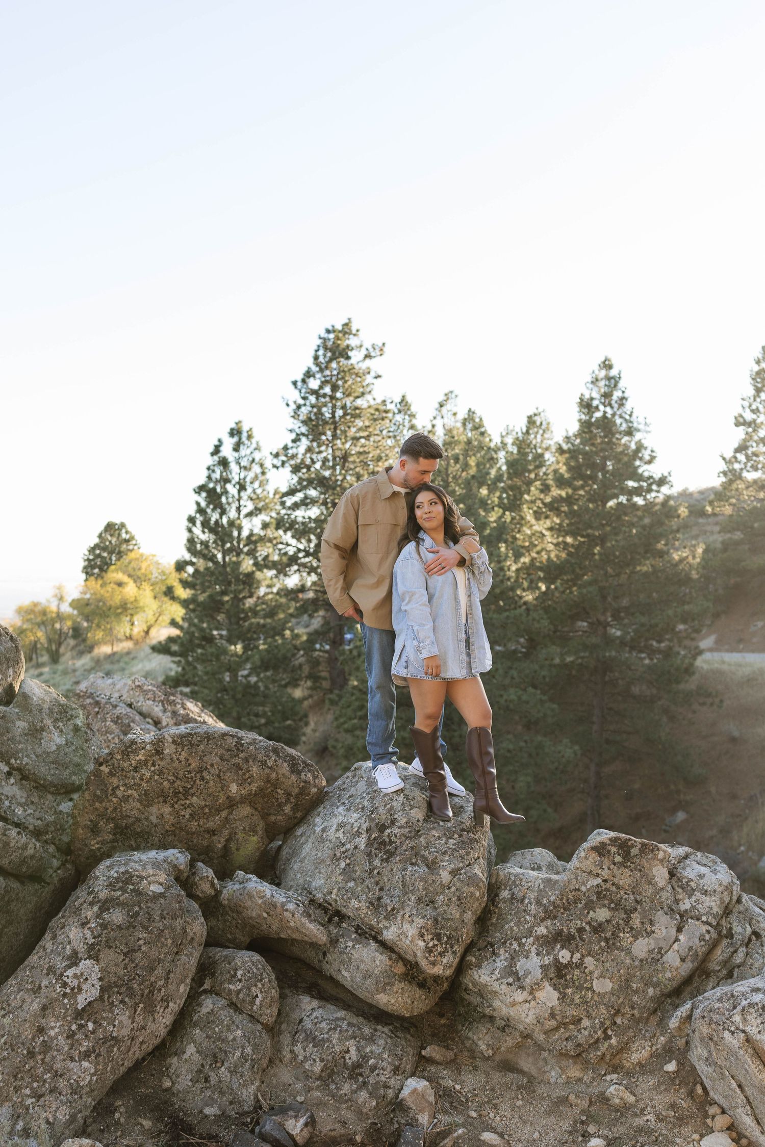 A couple stands together on a rocky outcrop surrounded by pine trees during a golden sunset photo session.