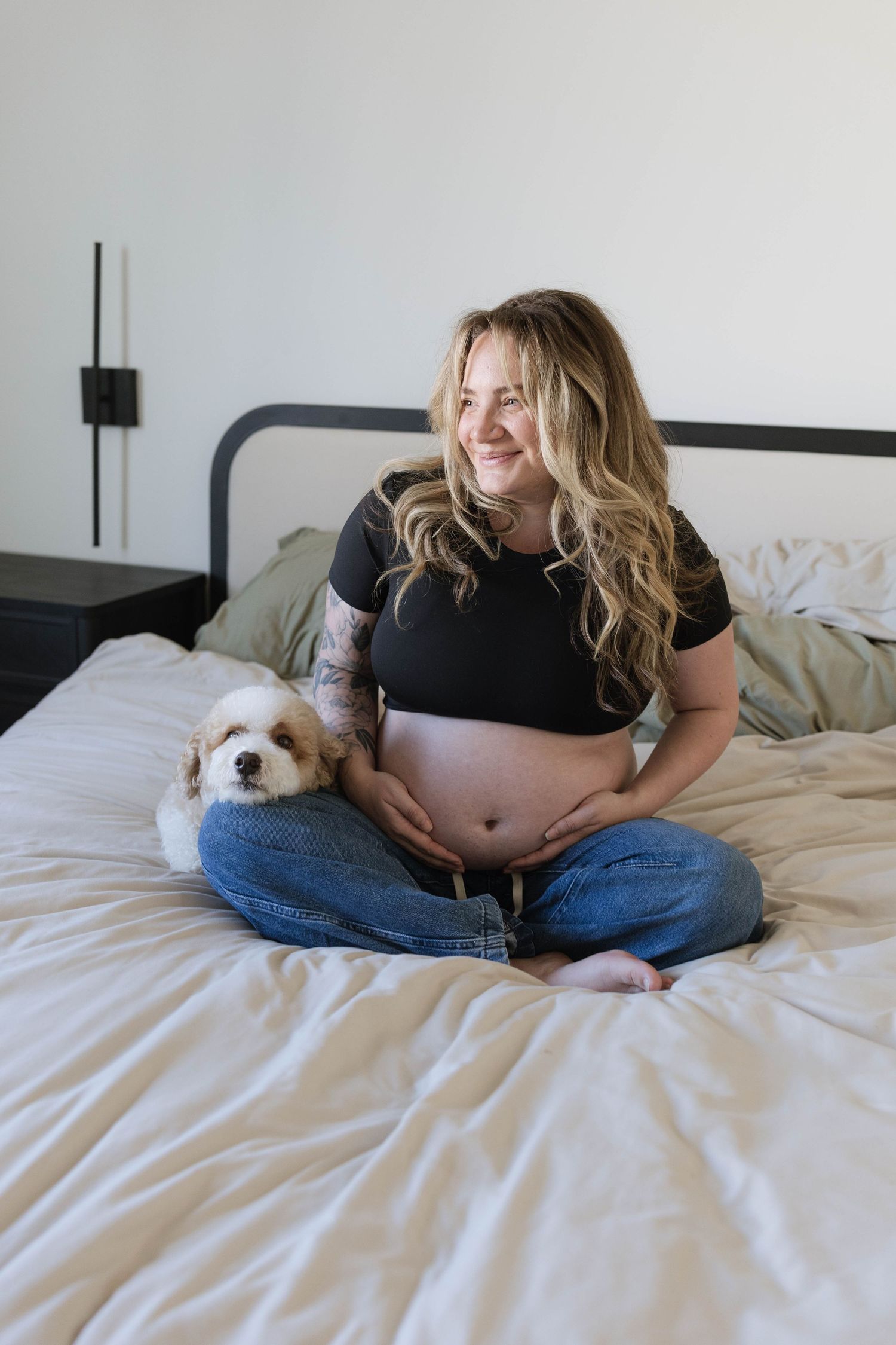 An expectant mother sits cross-legged on bed with small white dog.