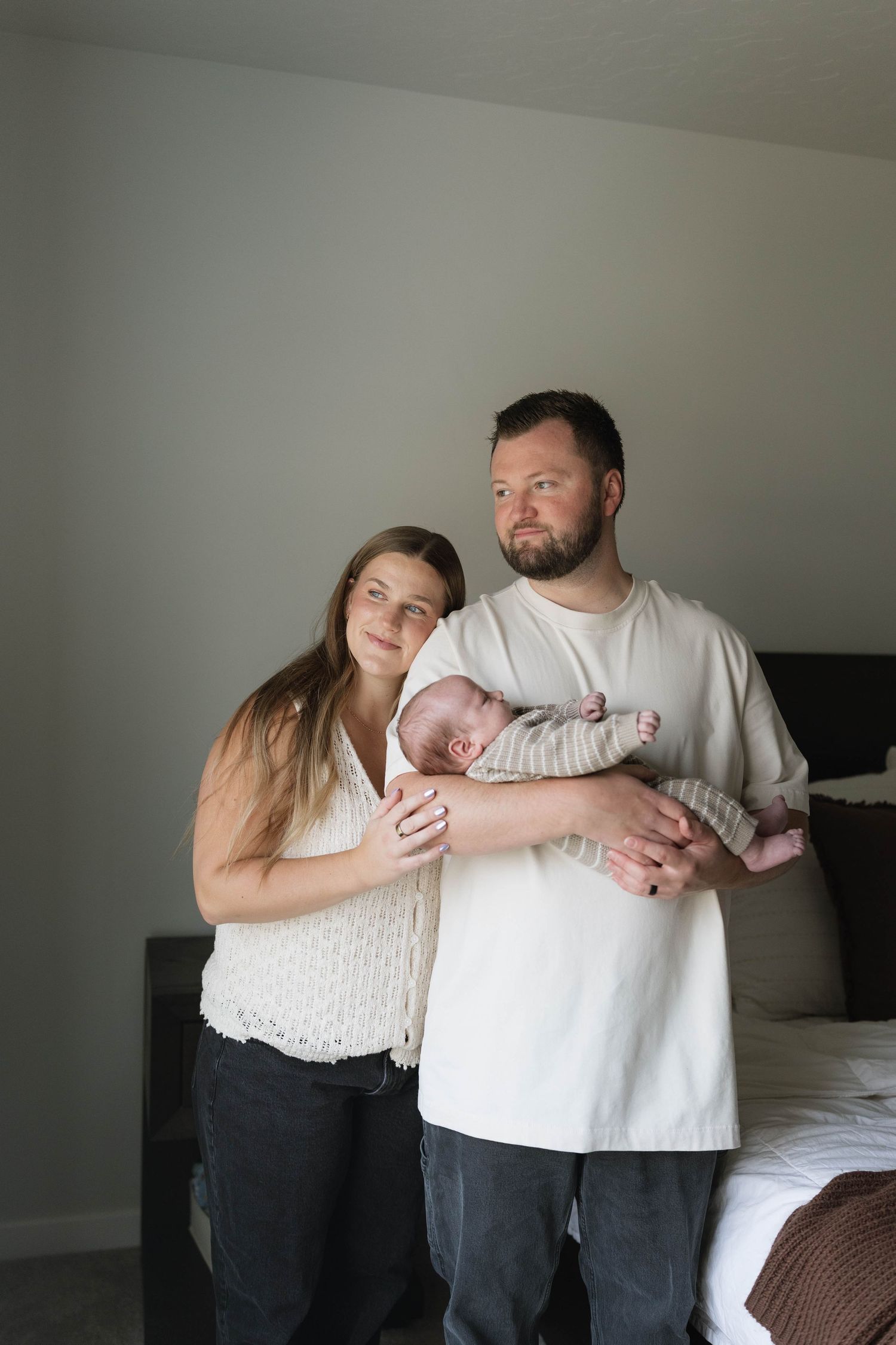 A couple stands together in a bedroom, with the man cradling a newborn baby while the woman rests her hand on his arm.