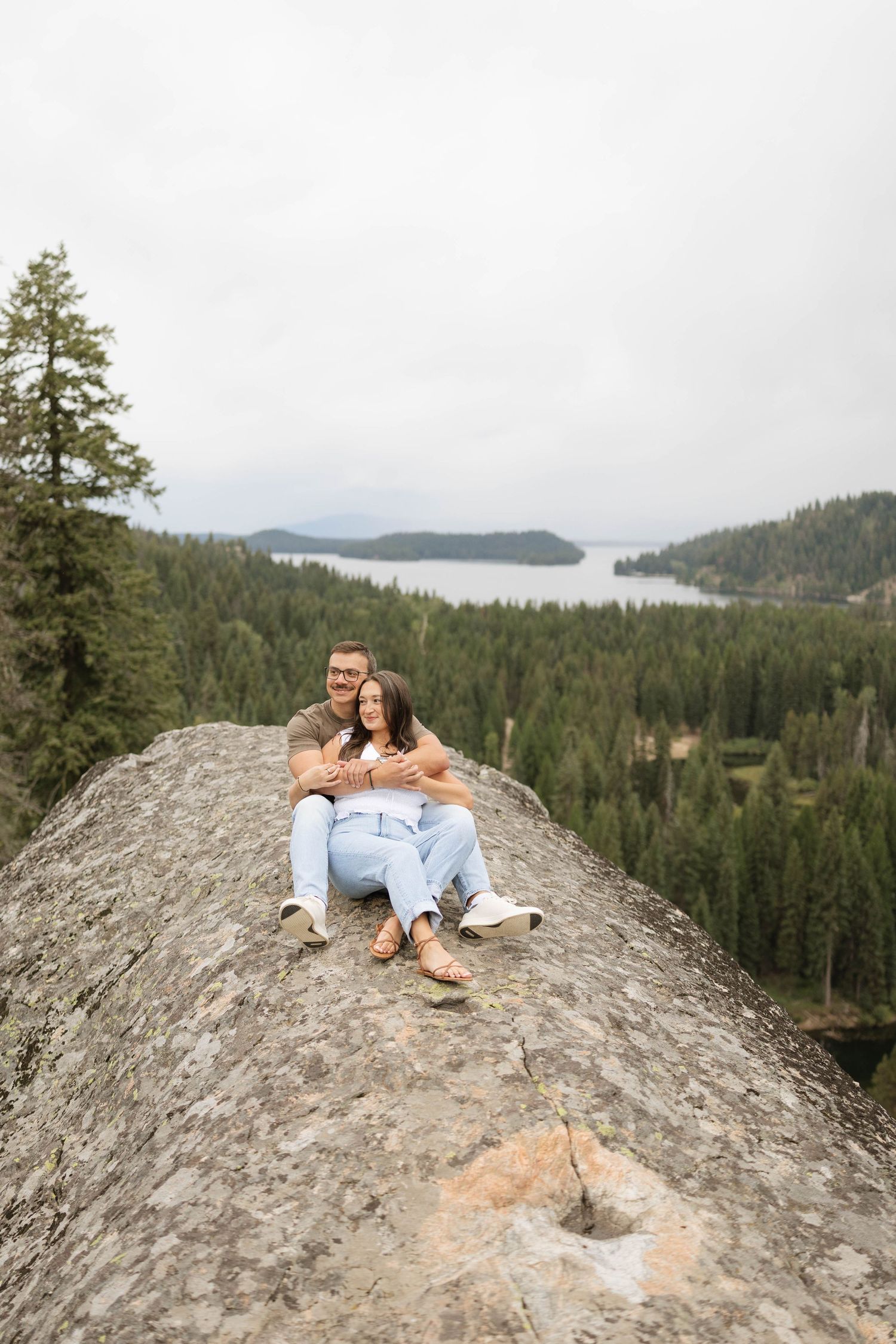 Couple sitting close together on a large granite rock overlooking a forested lake and distant hills under a cloudy sky.