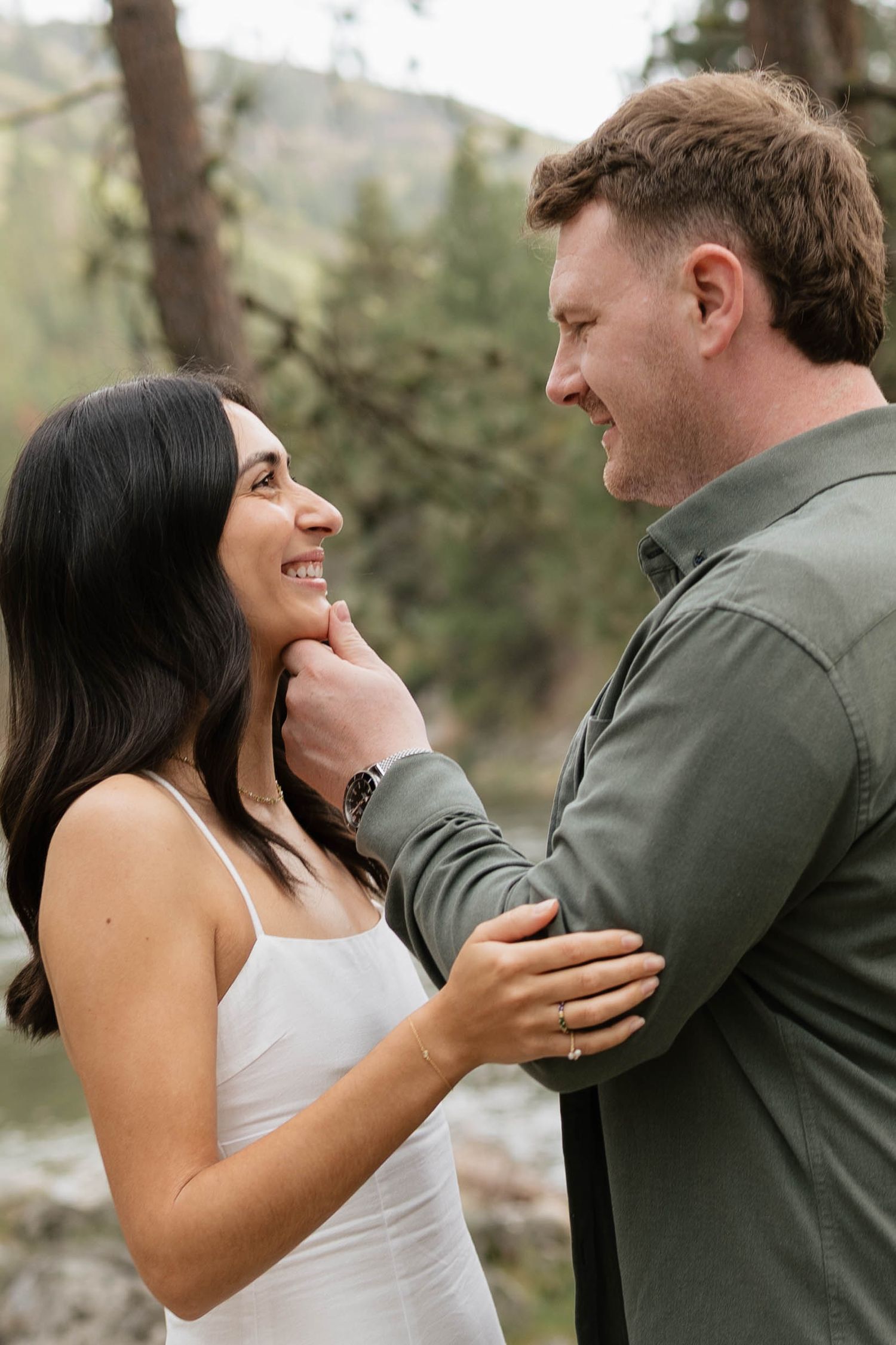 Couple sharing a tender moment outdoors, man gently touching woman's chin, smiling at each other in a forest setting.