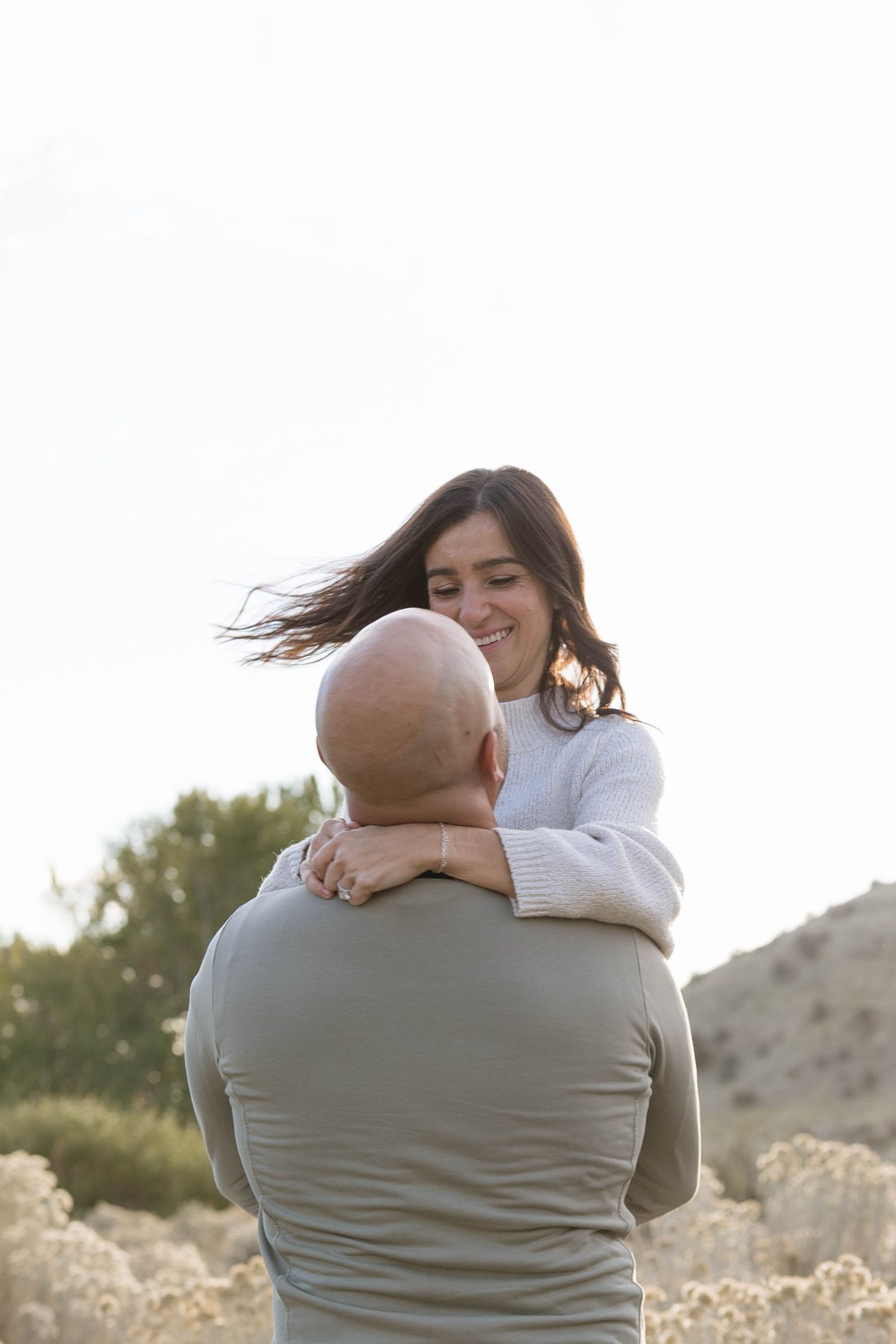 A couple embraces outdoors in a desert landscape with mountains in the background during golden hour.